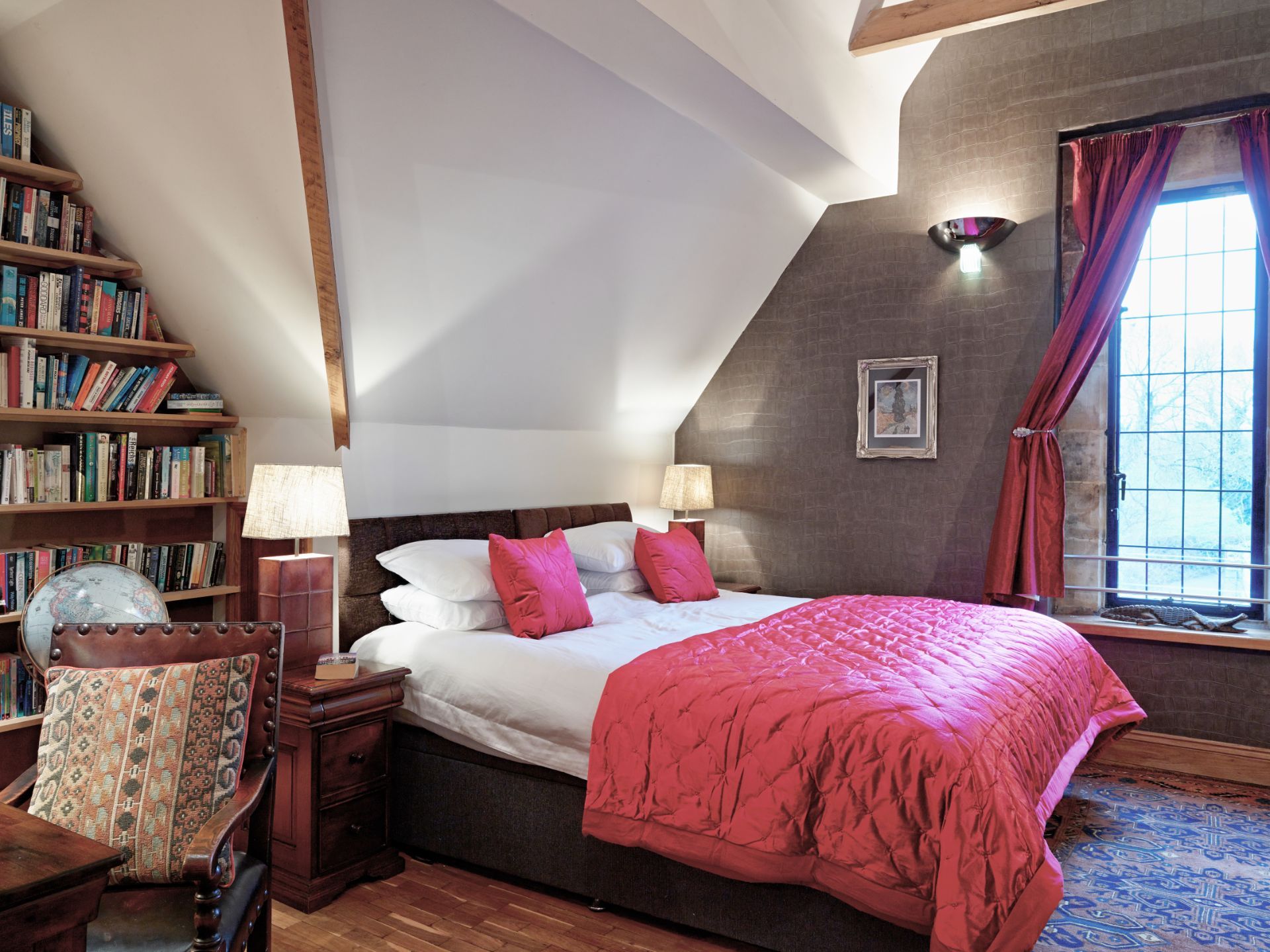 Cozy bedroom with a red bedspread, pink pillows, bookshelf, lamp, and window with curtains