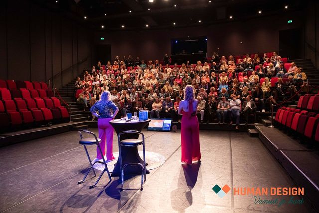 Twee vrouwen staan โโop het podium voor een groot publiek in een theater. De ene vrouw draagt โโeen roze jurk, de andere een rode.