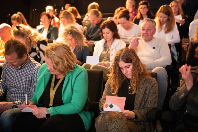 Toeschouwers zitten en maken aantekeningen tijdens een evenement. Mensen schrijven, geconcentreerd. Gedempte verlichting, binnenruimte.