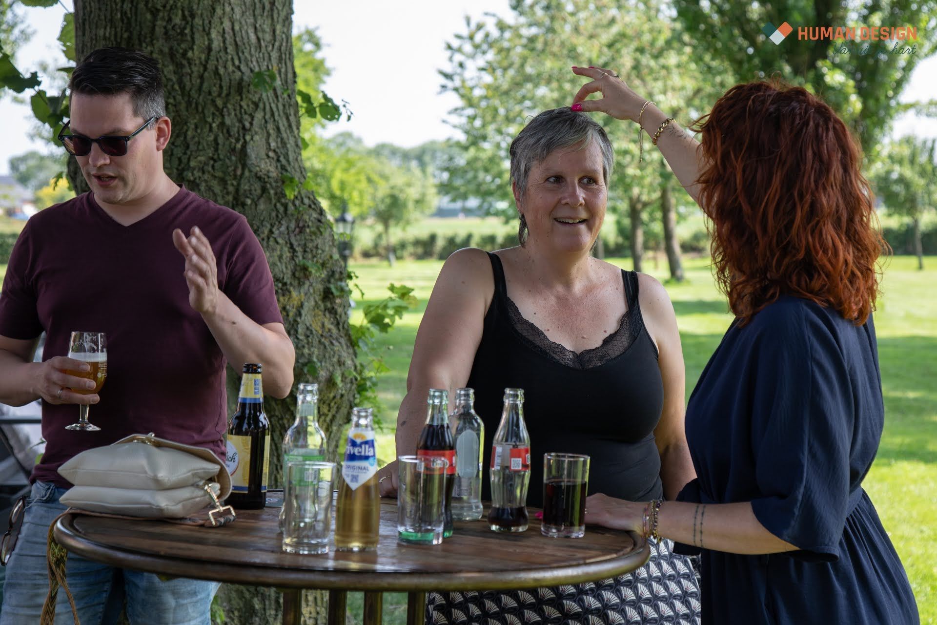 Mensen bij een bijeenkomst in de buitenlucht; een vrouw raakt het haar van een andere vrouw aan, drankjes op een tafel, zonnige dag.