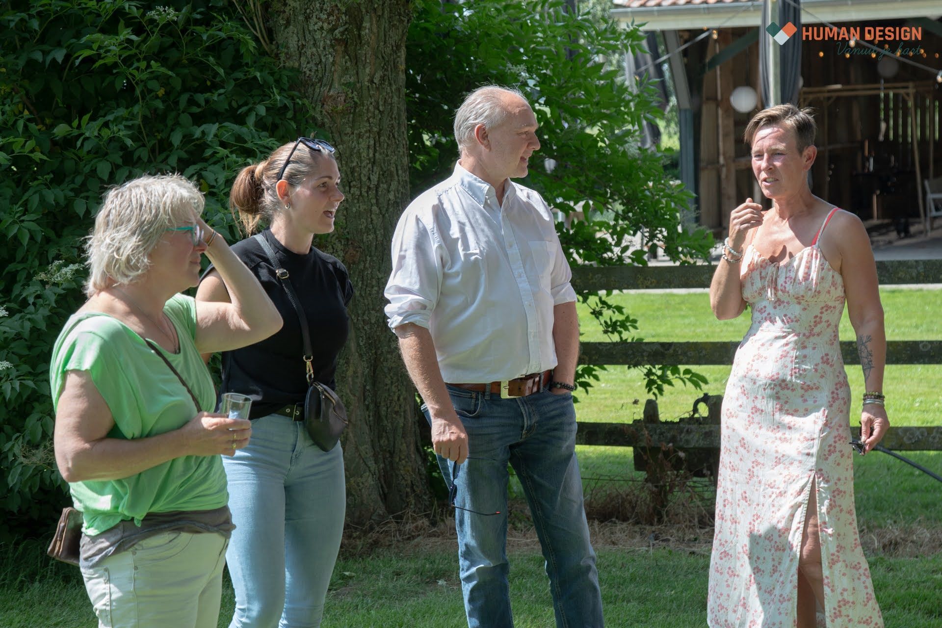 Vier mensen staan ββbuiten; een vrouw in een bloemenjurk spreekt. Anderen kijken toe, sommigen met een drankje in de hand, met bomen en een hek op de achtergrond.