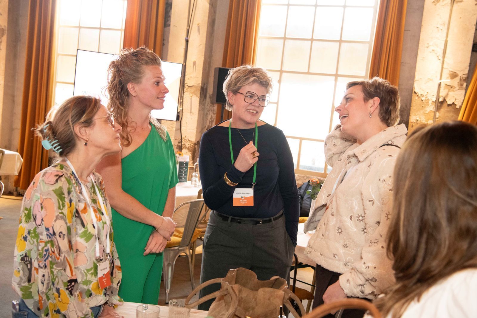 Drie vrouwen in een kamer, twee staan, één zit. Ze glimlachen en zijn in gesprek. Neutrale achtergrond, gedempt licht.