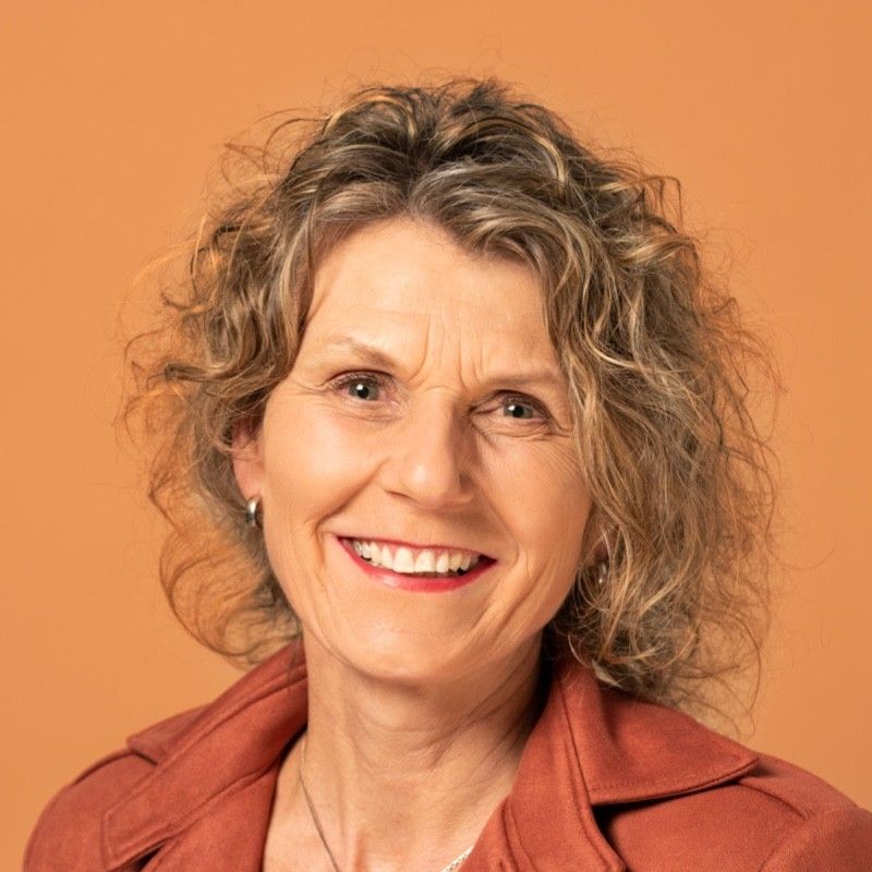 Een vrouw met krullend haar glimlacht, gekleed in een roestbruine jas, tegen een oranje achtergrond.