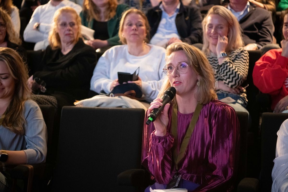 Vrouw die in een microfoon spreekt, te midden van een publiek, gekleed in een paarse blouse en met een bril op.