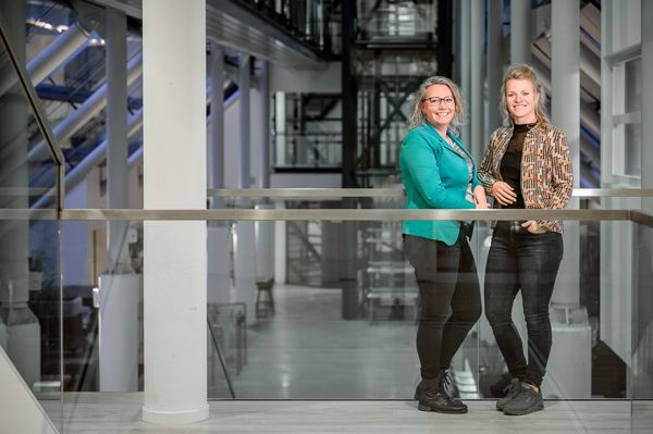 Twee vrouwen glimlachen en leunen tegen een reling in een gebouw met een industriële uitstraling. De ene draagt een turquoise jurk, de andere een jasje met een patroon.