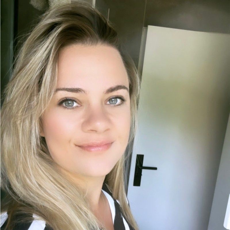 Een blonde vrouw glimlacht, van hoofd tot schouders, voor een witte deur.