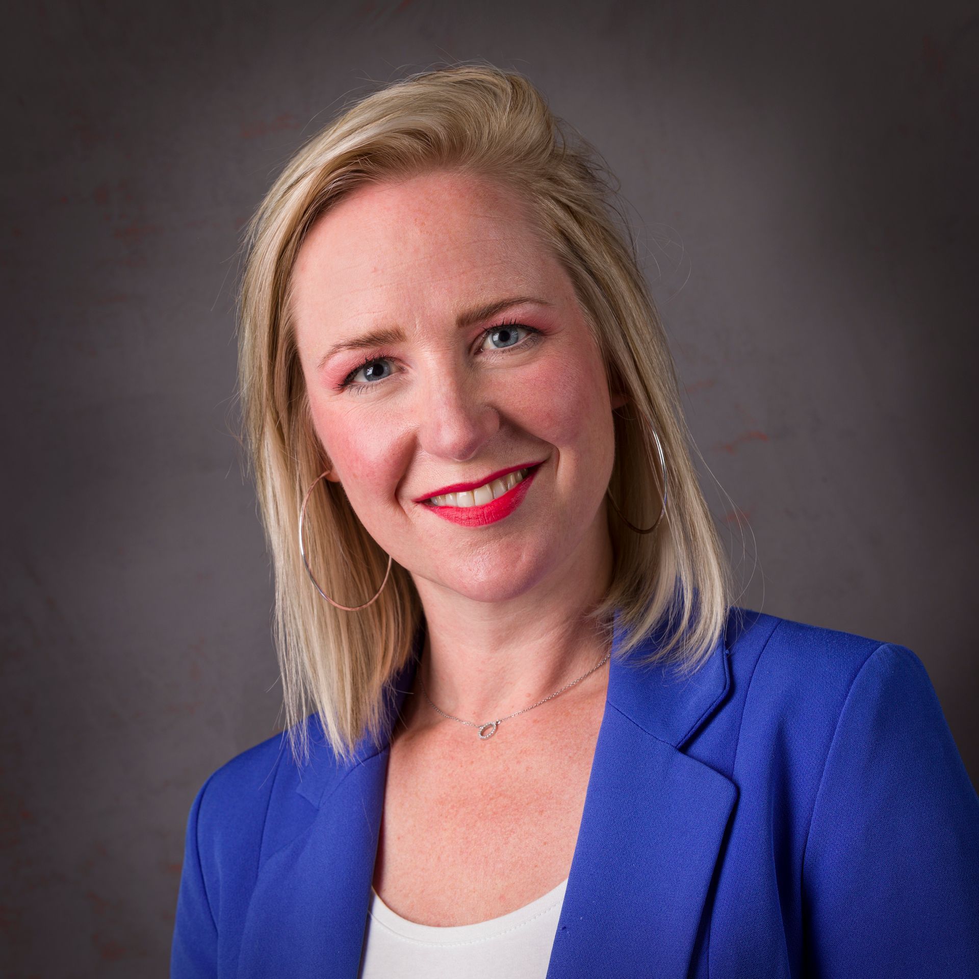 Een vrouw met blond haar, gekleed in een blauwe blazer en een witte top, lacht tegen een grijze achtergrond.