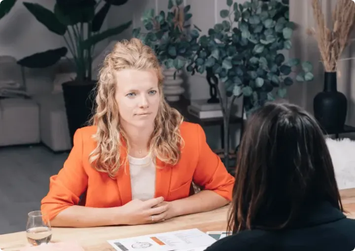 Twee mensen zitten tegenover elkaar aan een tafel in een vergadering; een van hen draagt ​​een oranje colbert en spreekt met documenten tussen zich in.