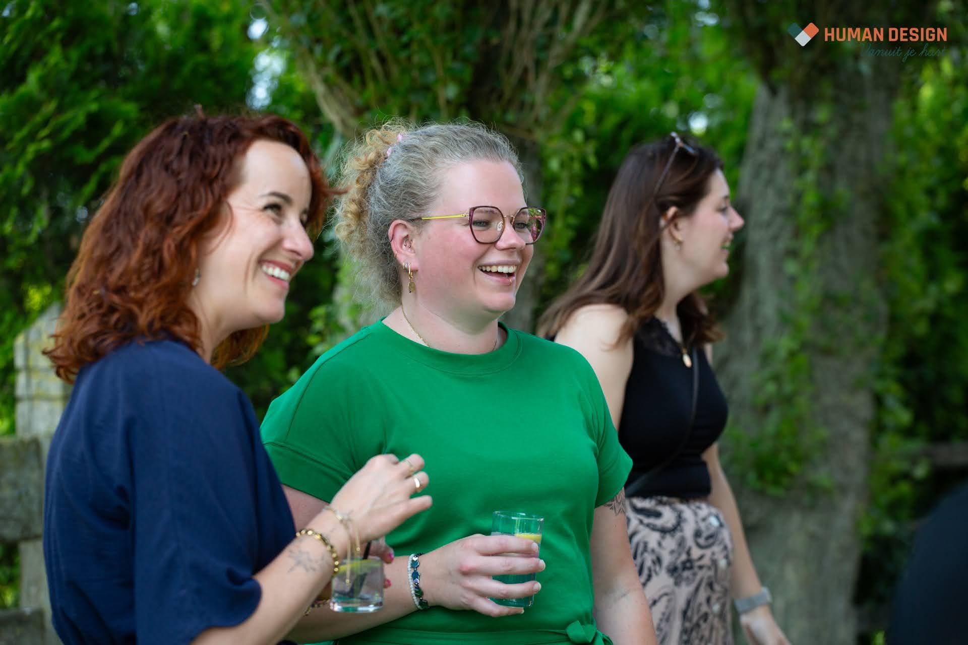 Drie lachende mensen buiten; een vrouw in een groen shirt met een drankje in haar hand, en andere vrouwen die ook lachen.