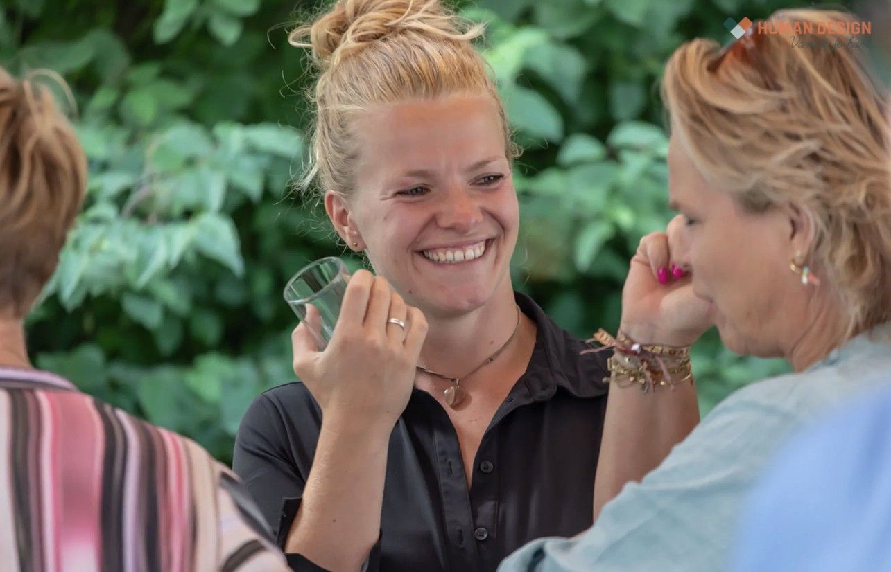 Een vrouw lacht, houdt een glas vast en praat met anderen buiten.