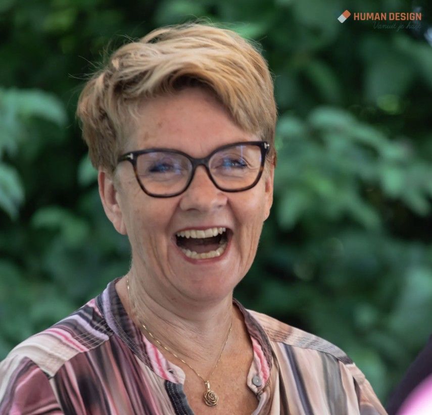 Een vrouw met kort blond haar en een bril lacht, terwijl ze een gestreept shirt draagt ββen buiten staat.