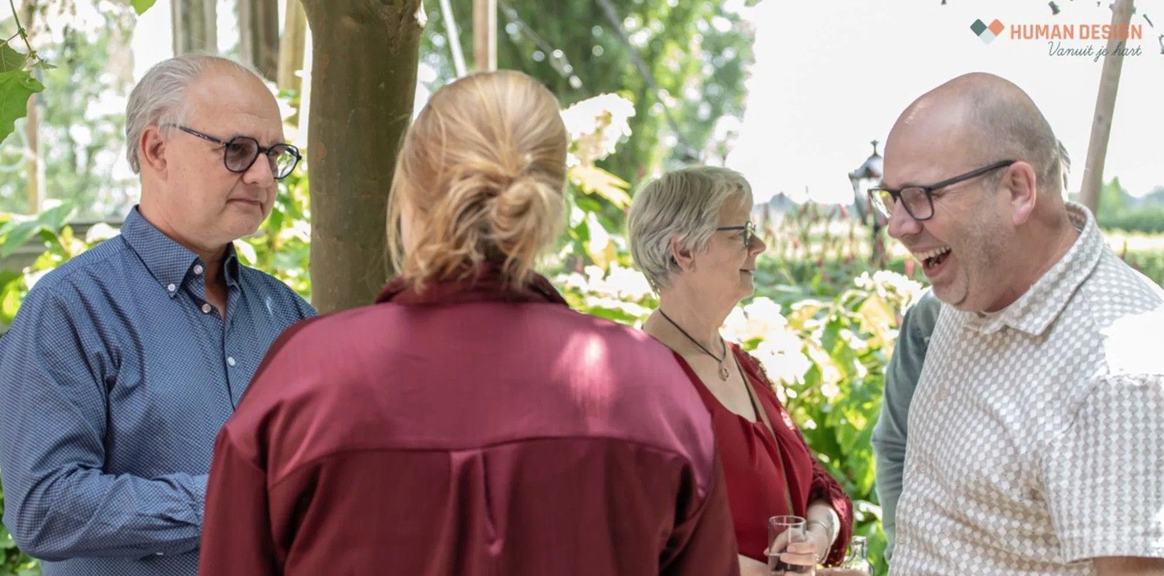 Vier mensen in gesprek in de buitenlucht, twee dragen een bril, glimlachen, groen op de achtergrond.