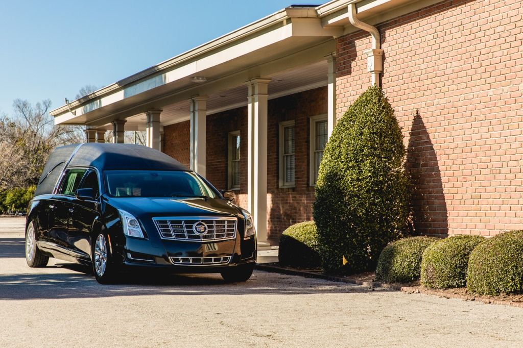 Black hearse parked in front of a brick building with a covered walkway and manicured shrubs.