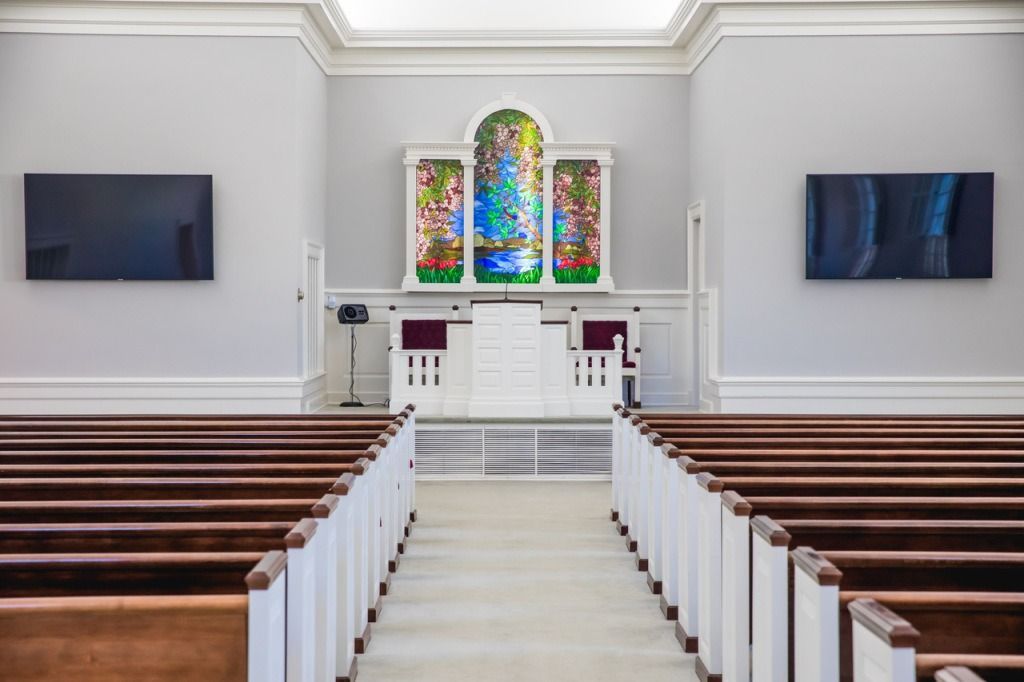 Rows of wooden pews face an altar with a stained glass panel, flanked by two mounted screens.
