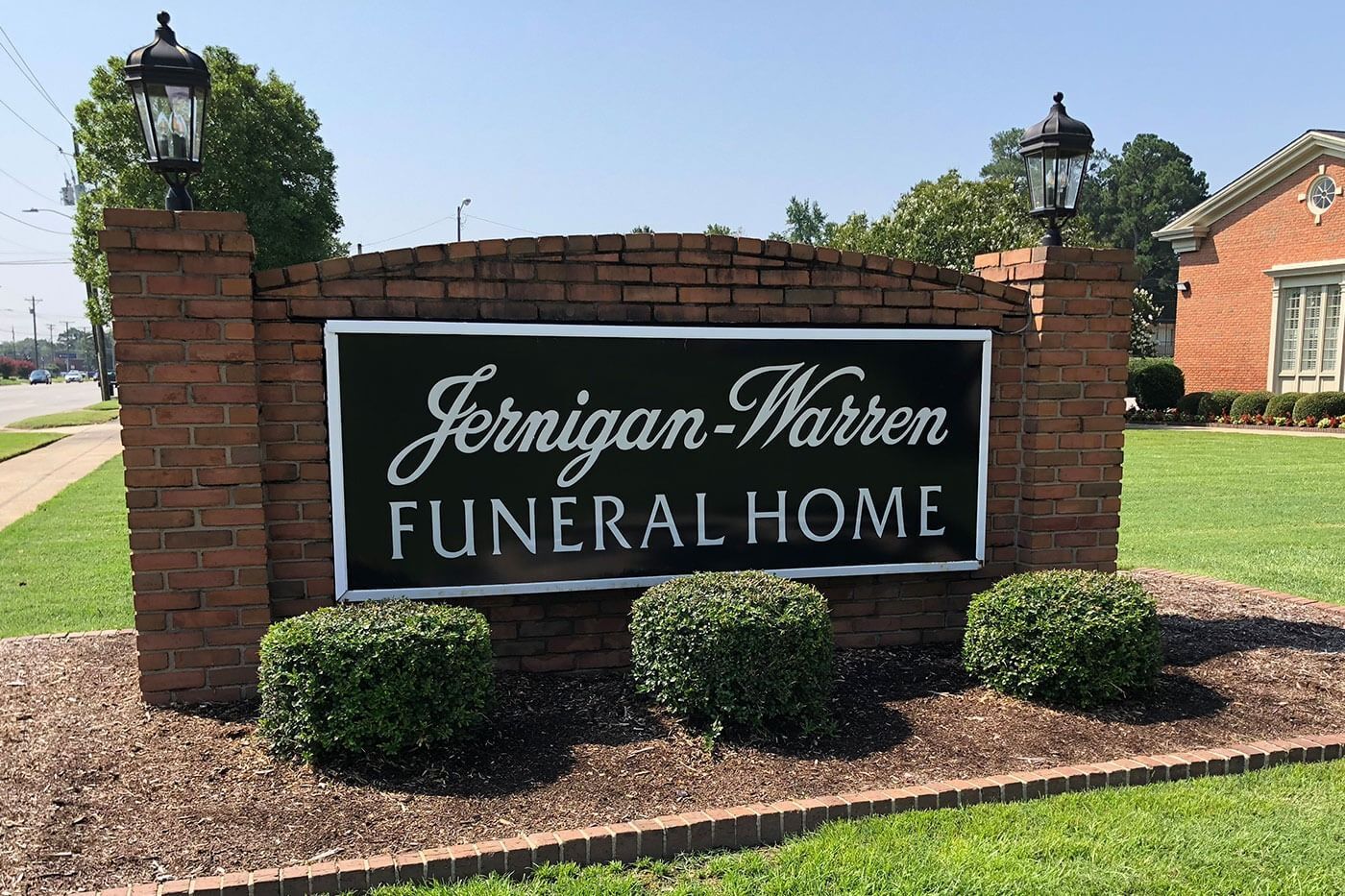 Sign for Jernigan-Warren Funeral Home, black text on a dark background within a brick structure, shrubbery in front.