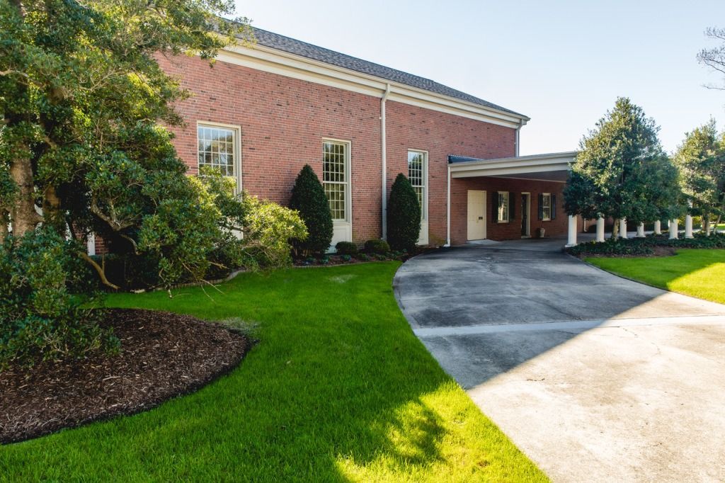 Brick building with arched windows, covered entrance, and curved driveway on a grassy lawn.