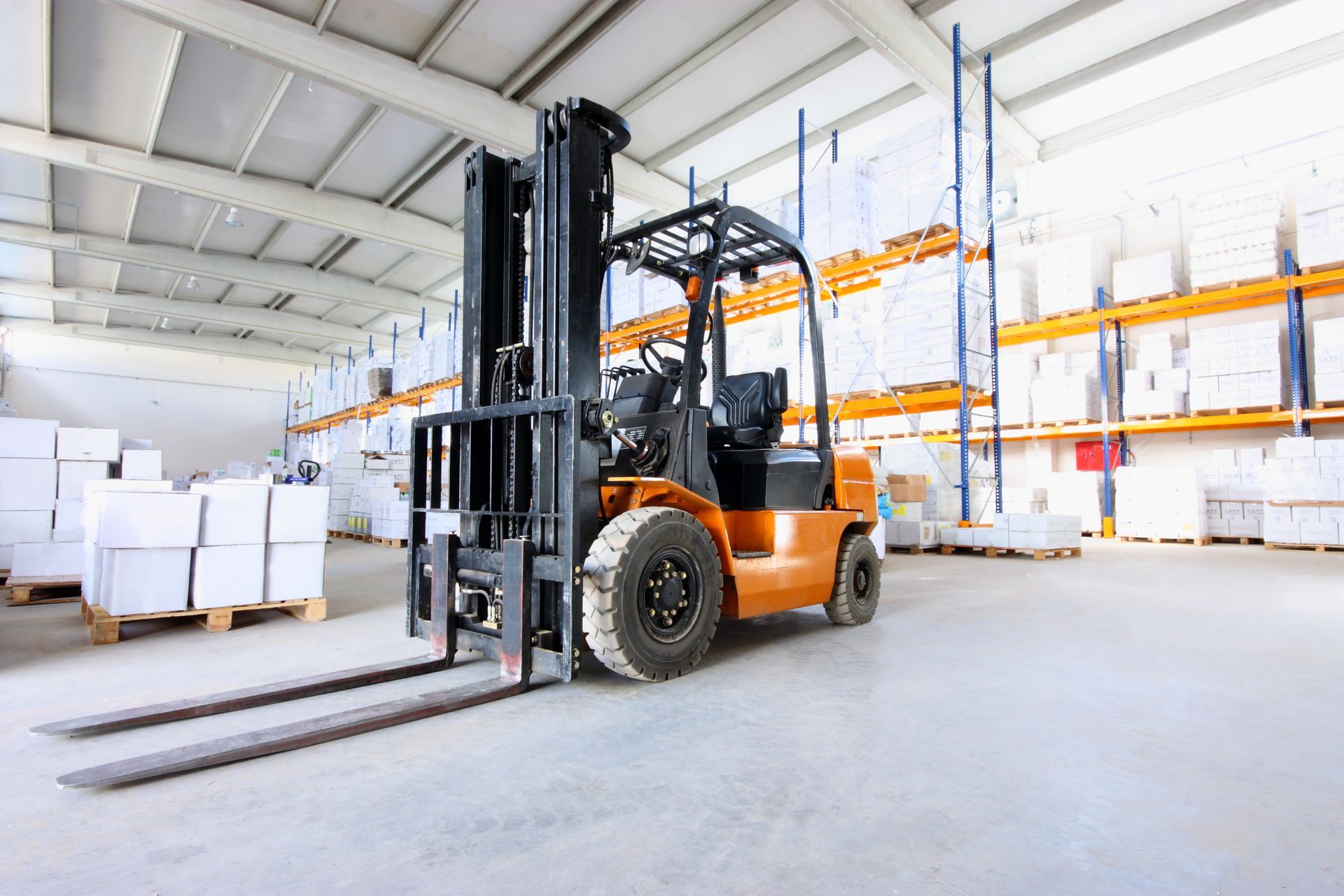 Orange forklift in a bright warehouse with stacked boxes and pallets