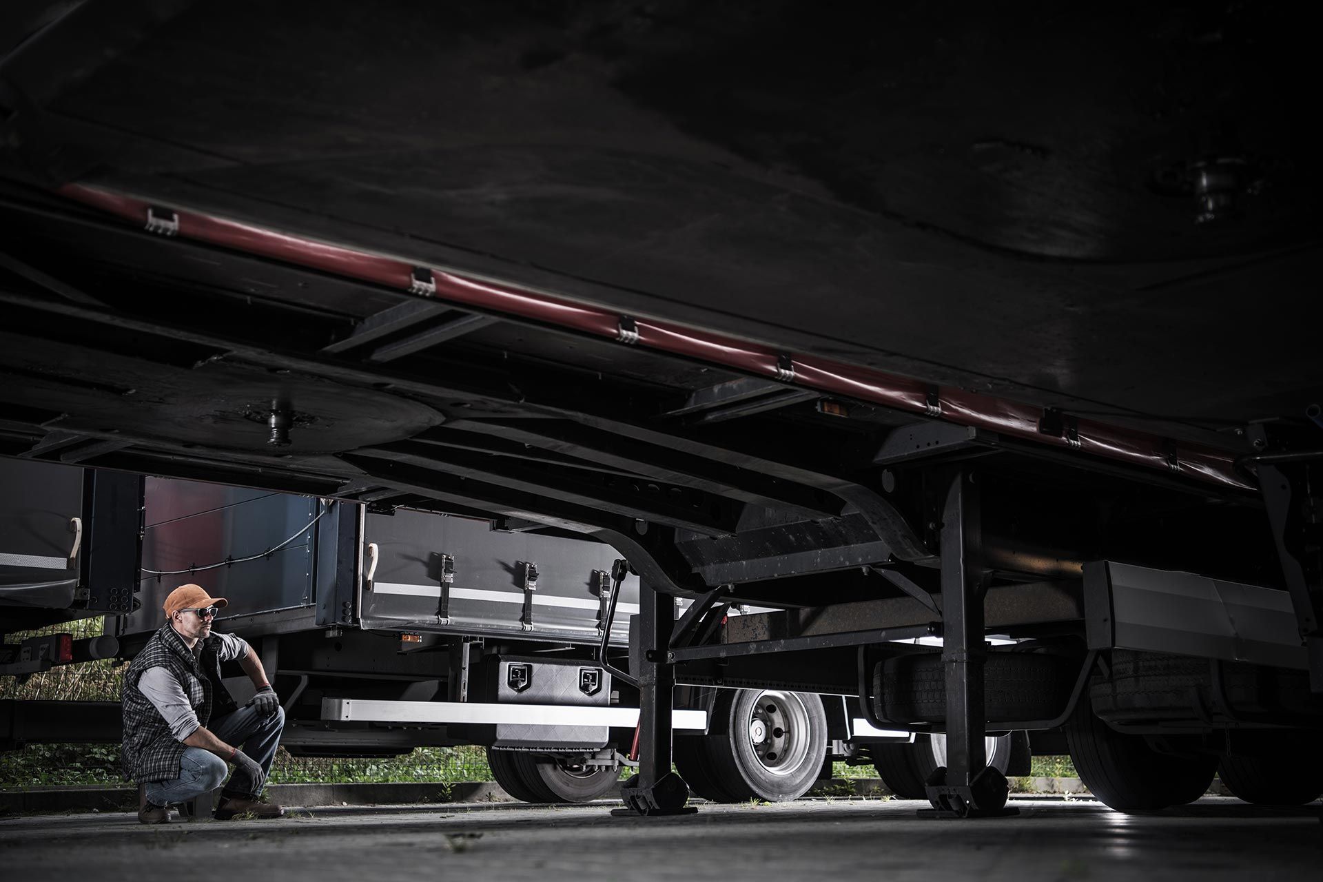 Person inspecting the underside of a parked semi-trailer in a loading area. Person inspecting the underside of a parked semi-trailer in a loading area.