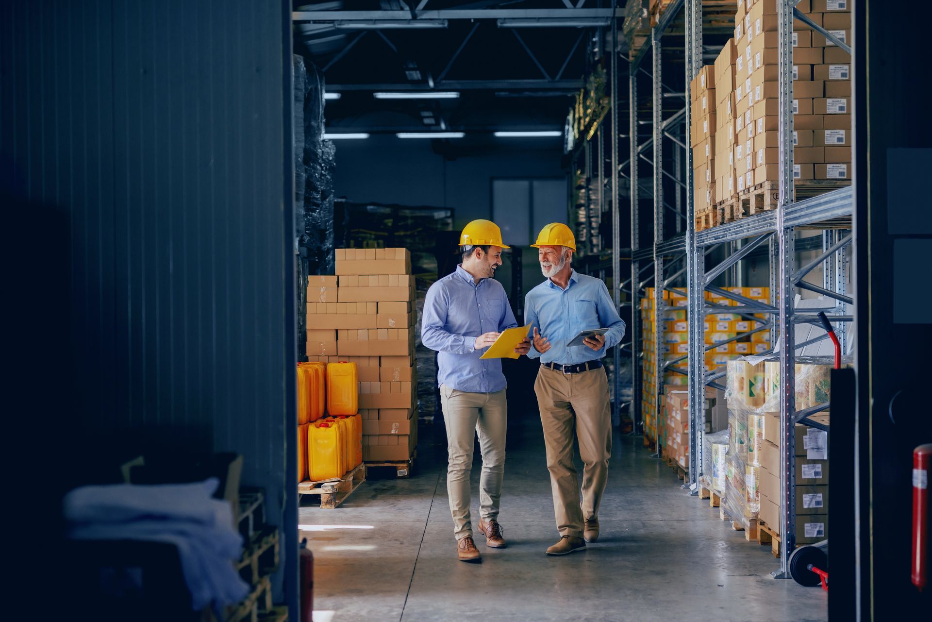 Two business partners in formal wear and helmets, from a logistics company, are in a warehouse. Two business partners in formal wear and helmets, from a logistics company, are in a warehouse.
