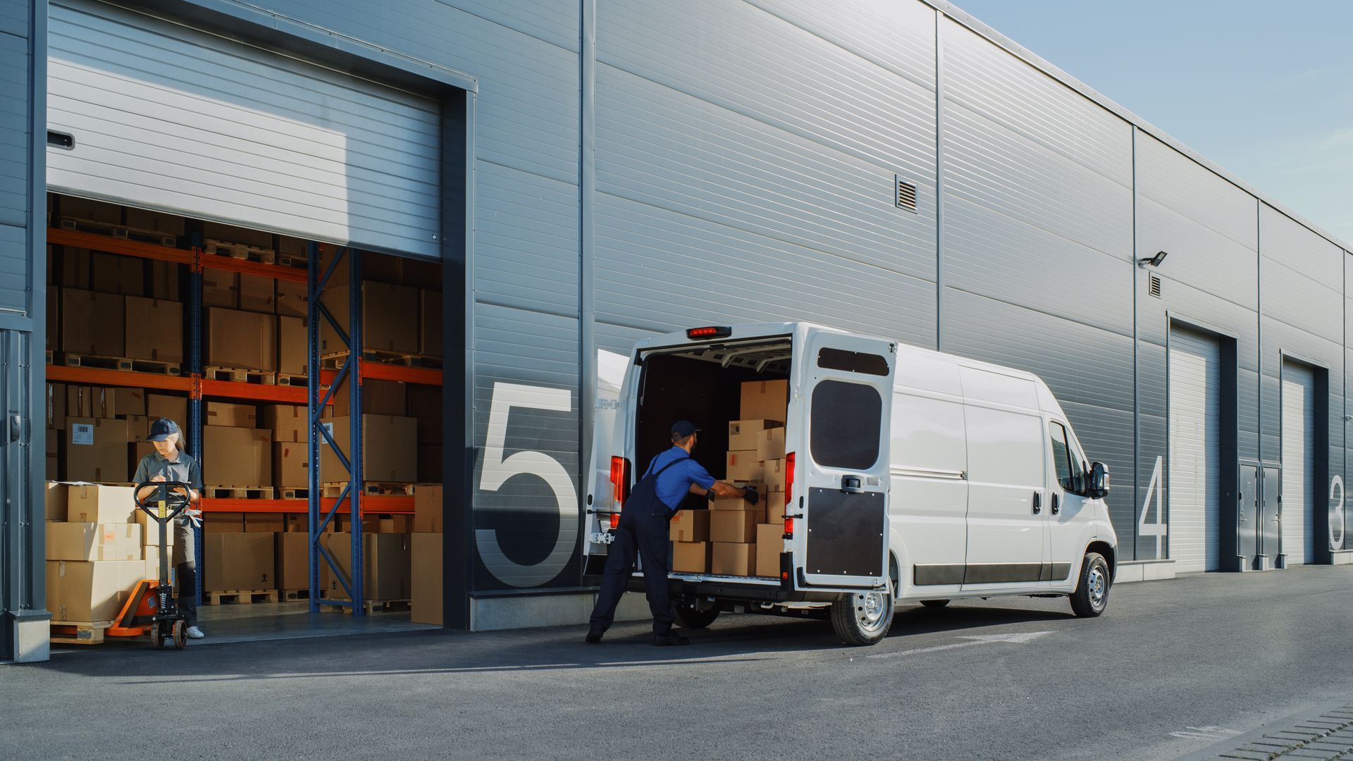 Worker loading boxes into a delivery van outside a warehouse with open loading bay doors.