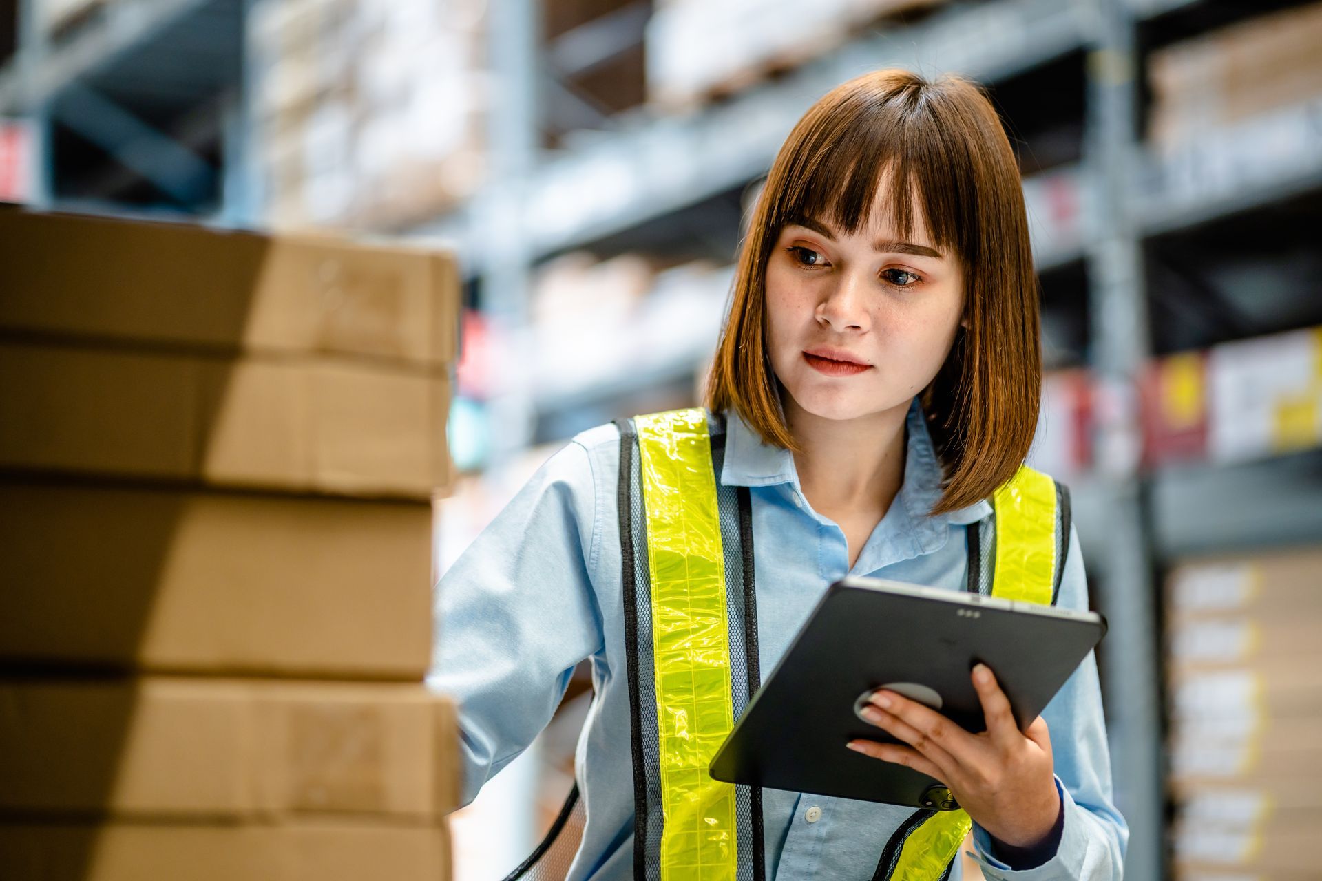 Warehouse worker using a tablet to manage inventory and inspect stacked boxes.