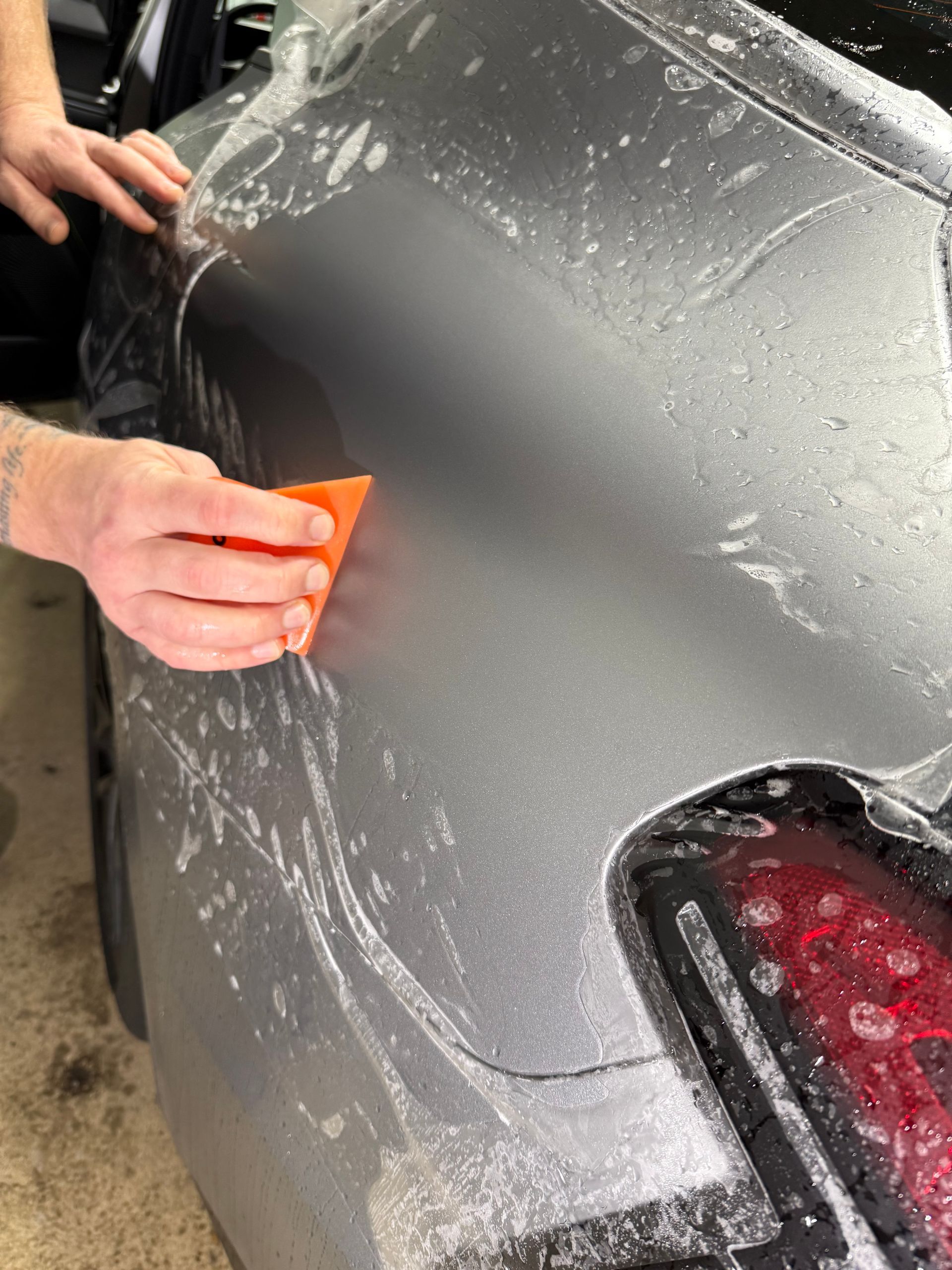 A person is using a blue squeegee to remove plastic from the hood of a car