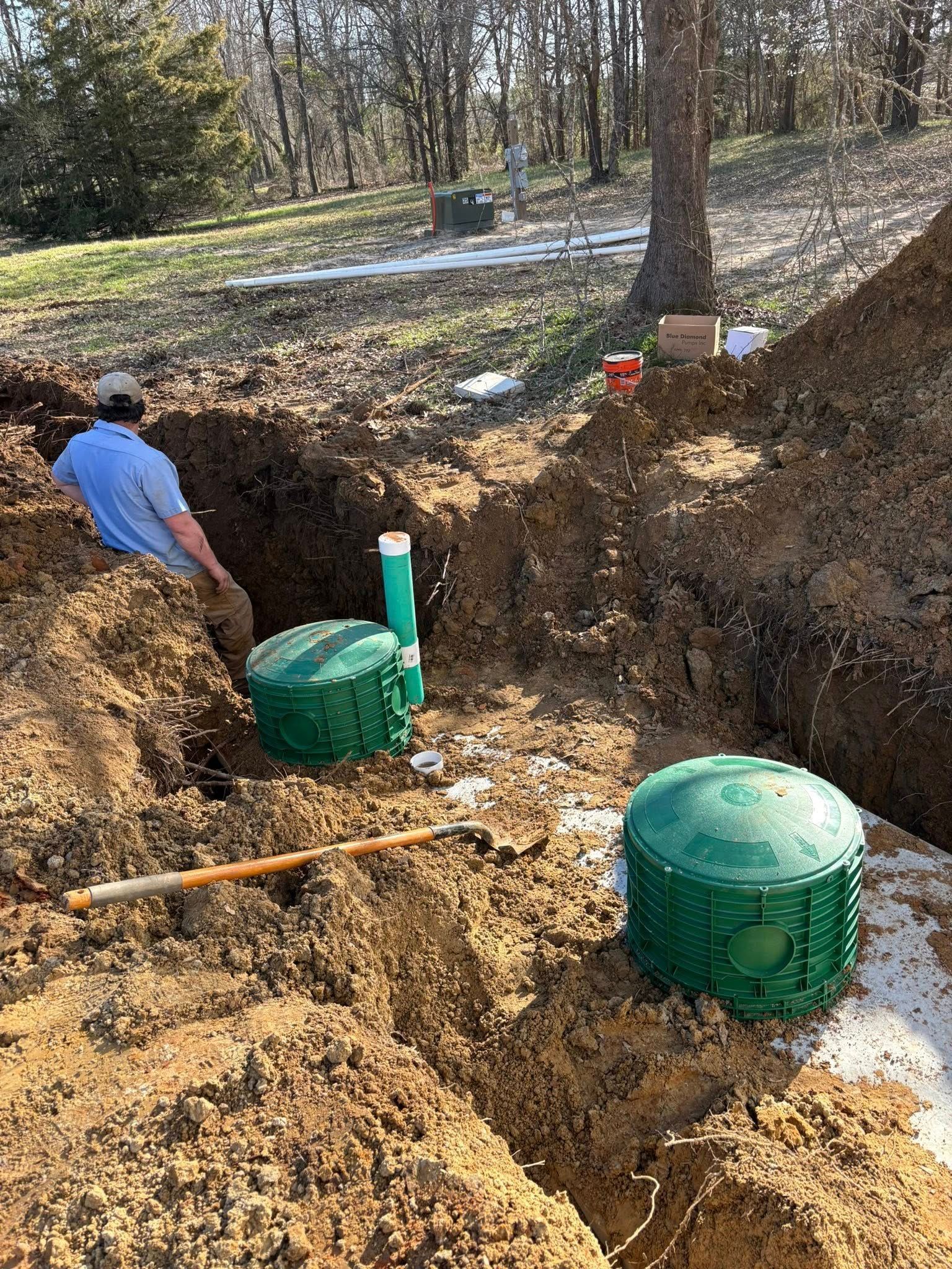 Man working on septic tank installation in a trench. Green tanks, soil, shovel, trees, outdoors.