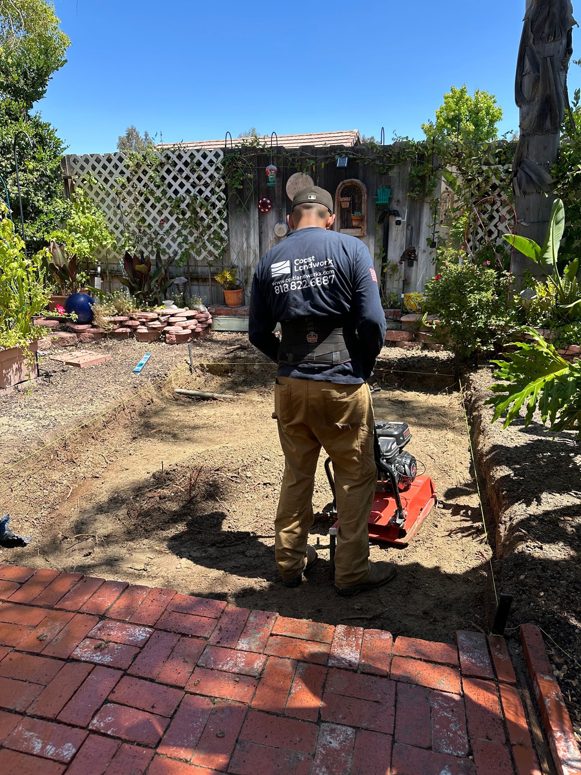 Worker using a leaf blower in a backyard garden with lattice fence and red patio bricks