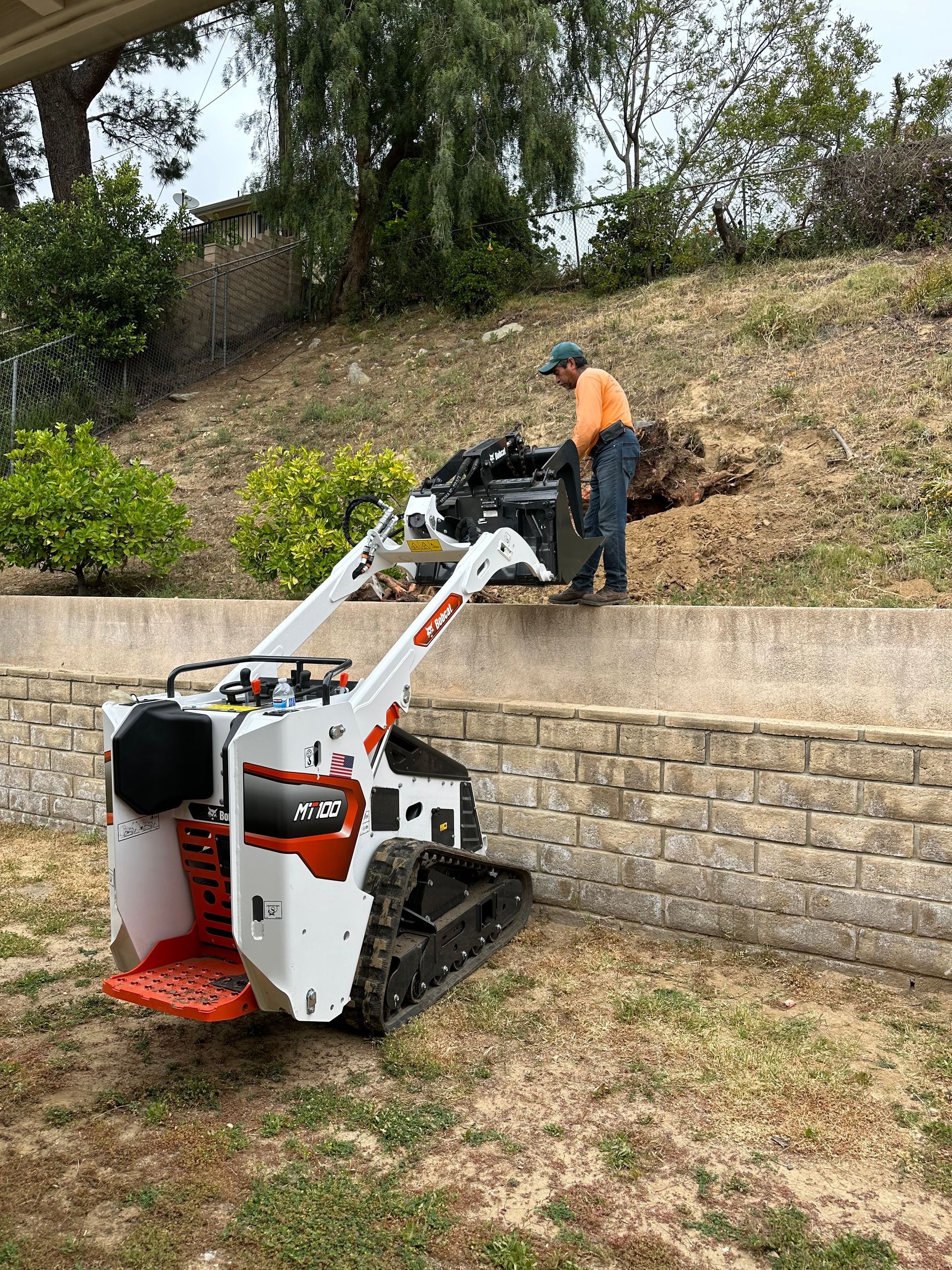 Compact loader lifting soil as a worker in an orange vest stands beside a stone retaining wall.