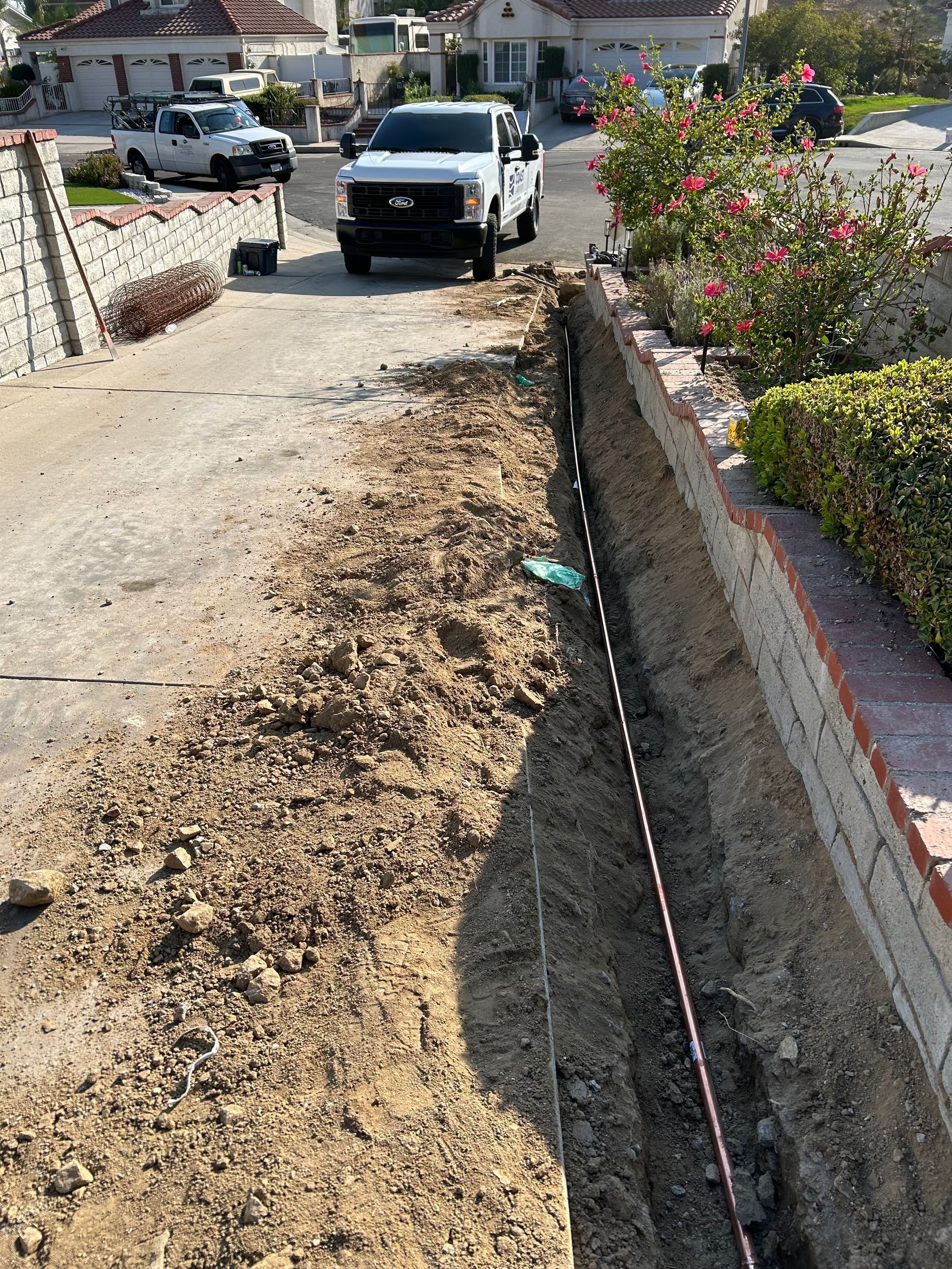 Roadside trench under construction beside a dirt lane, with a white pickup truck and houses in the background.