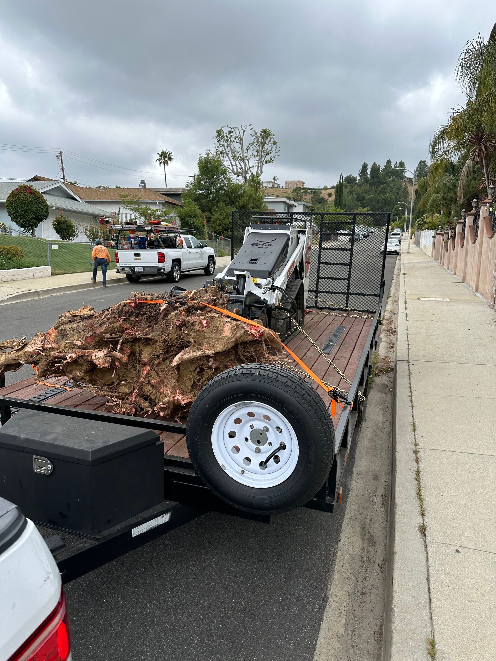 Debris-filled utility trailer parked on a street beside a sidewalk and fence after cleanup work