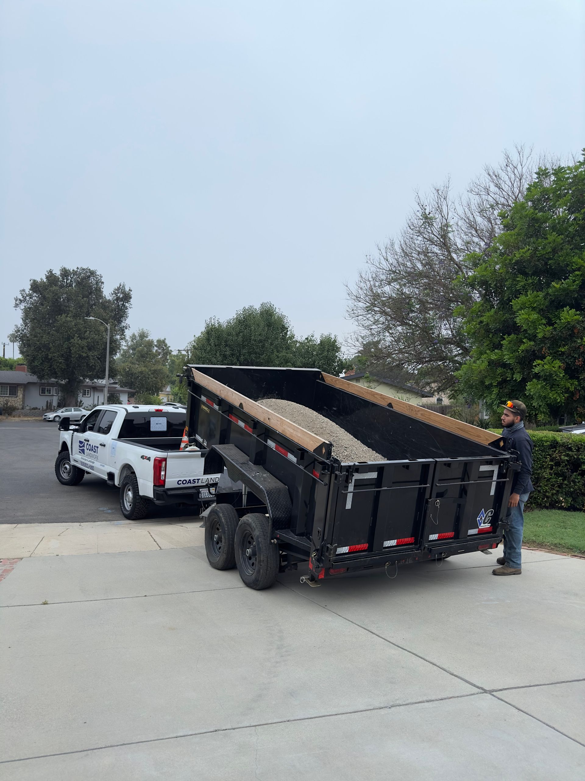 Worker unloading a dump trailer filled with mulch beside a white pickup on a driveway