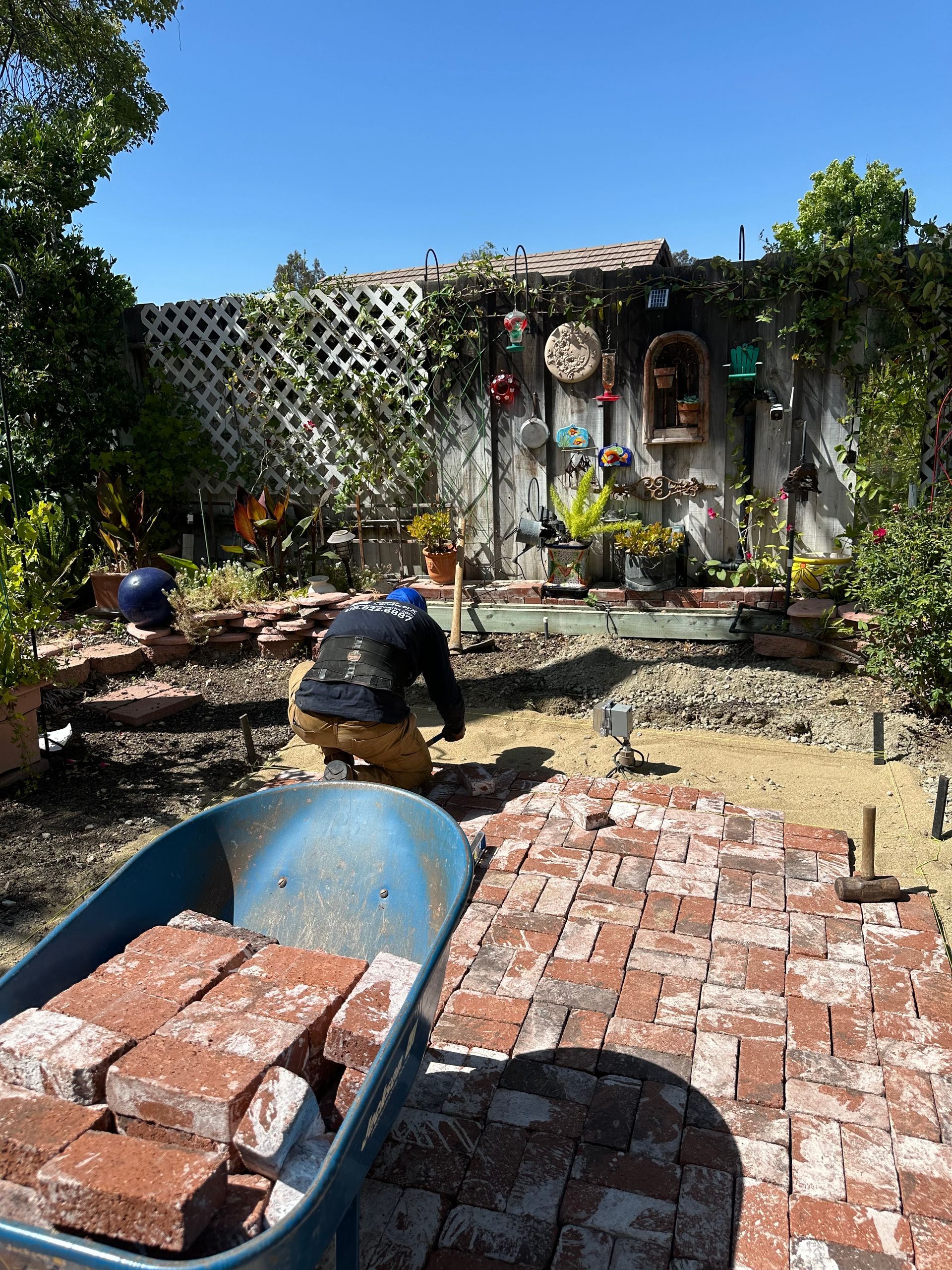 Person working in a sunlit backyard with bricks, a wheelbarrow, and garden decorations.