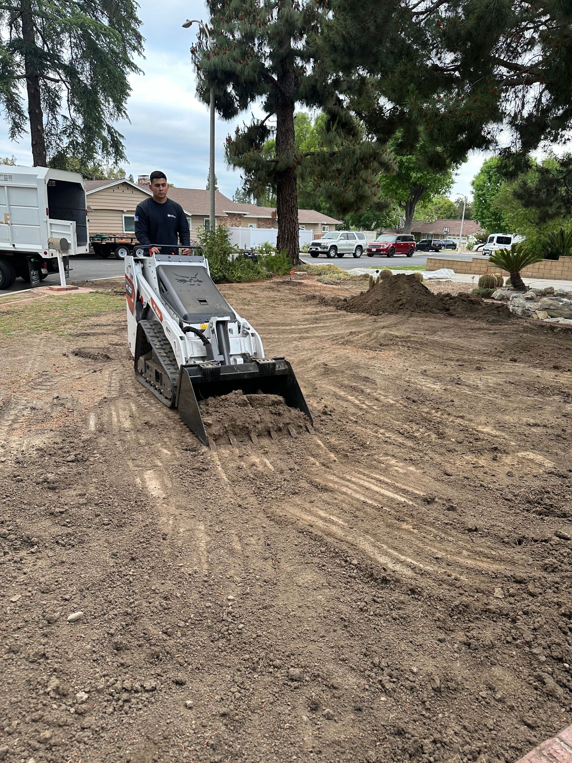Worker operating a compact loader on a dirt mound at a construction site with trees and trucks nearby