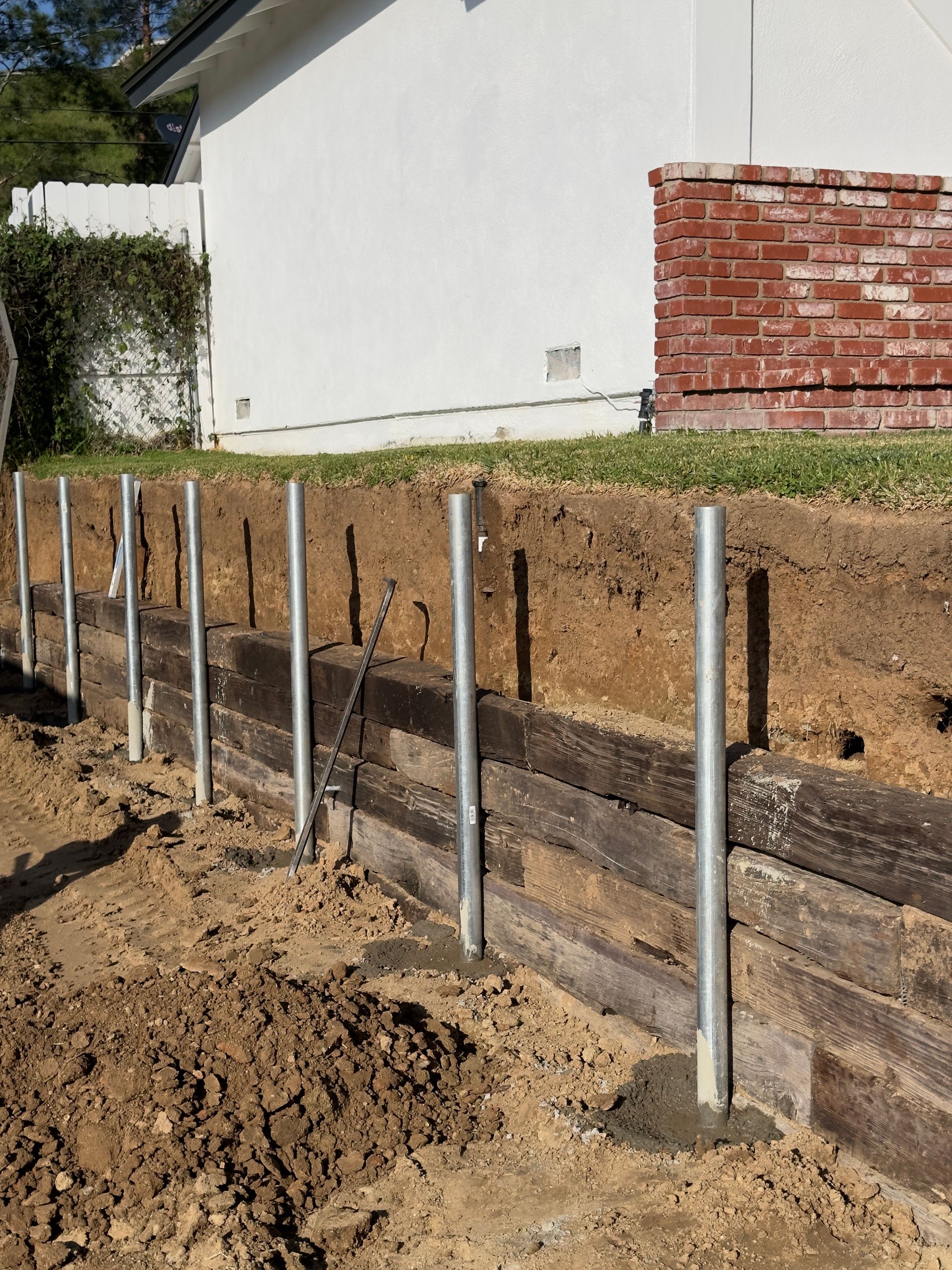 Construction site with steel posts set in concrete beside a retaining wall and a brick house.