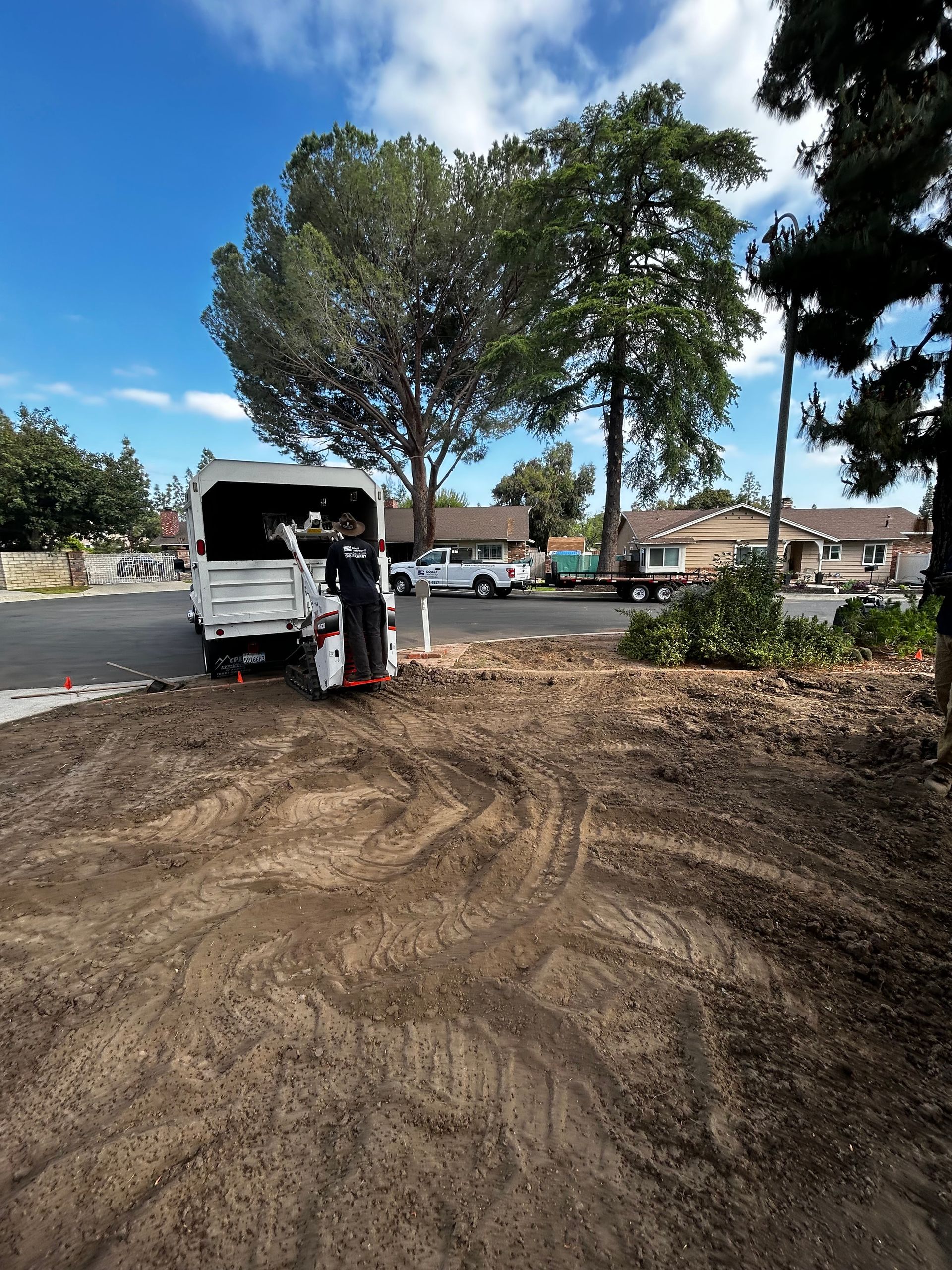 Worker unloading materials from a white truck on a dirt lot near houses and trees under a blue sky