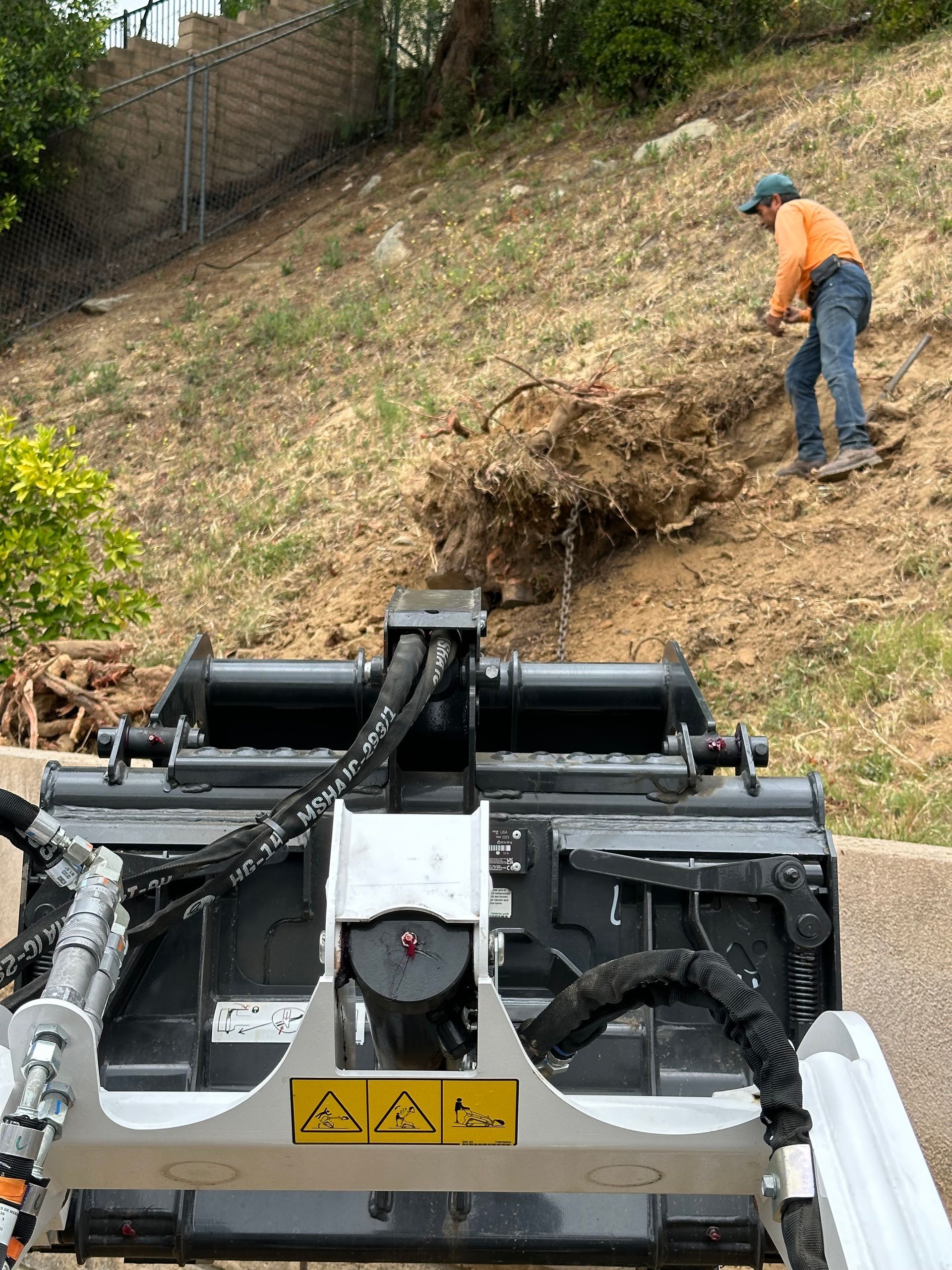 Worker in orange shirt clearing a large brush pile on a steep hillside beside a machine in the foreground
