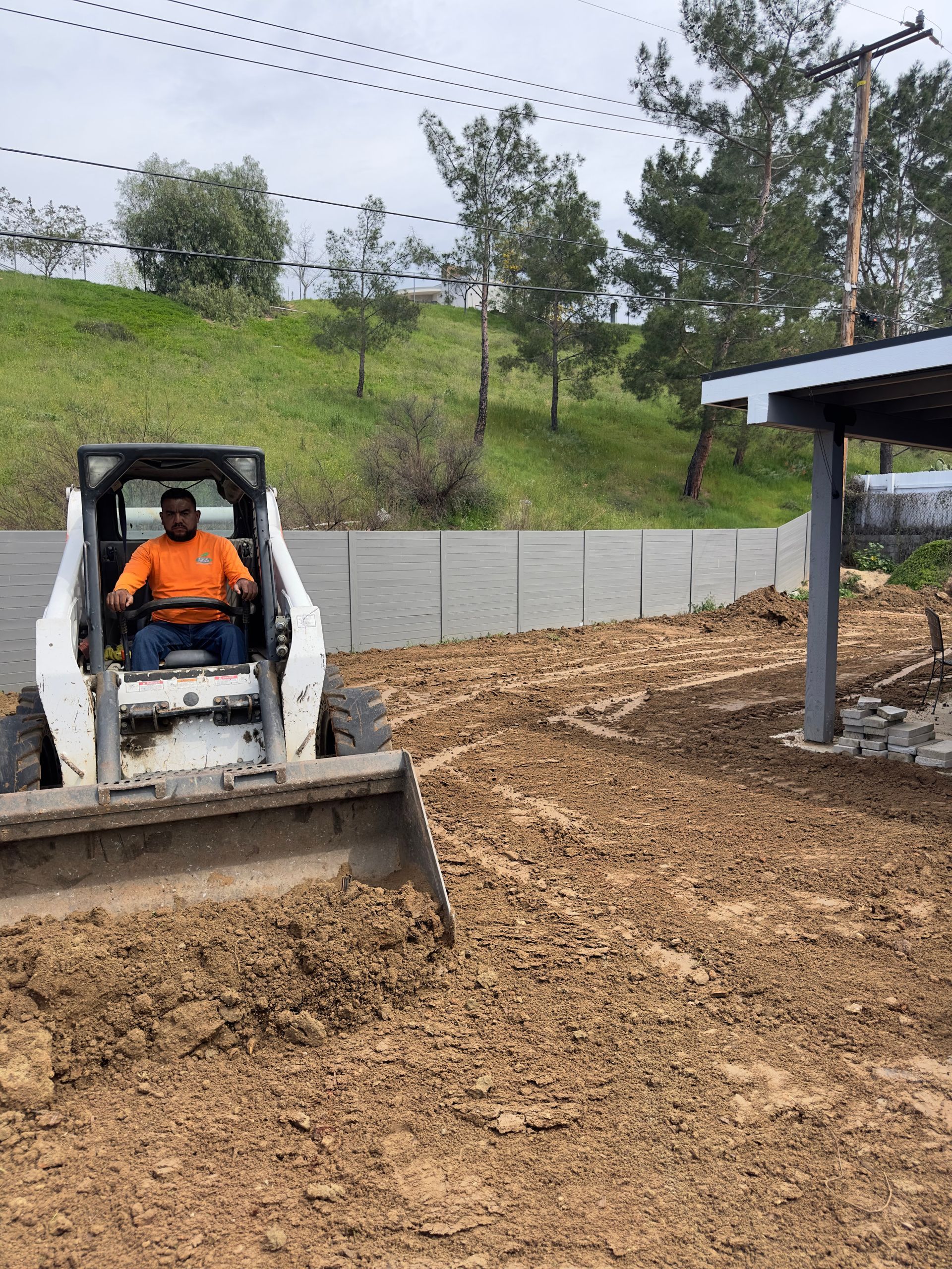 Small skid steer spreading dirt in a yard beside a covered structure, with a grassy hill behind.