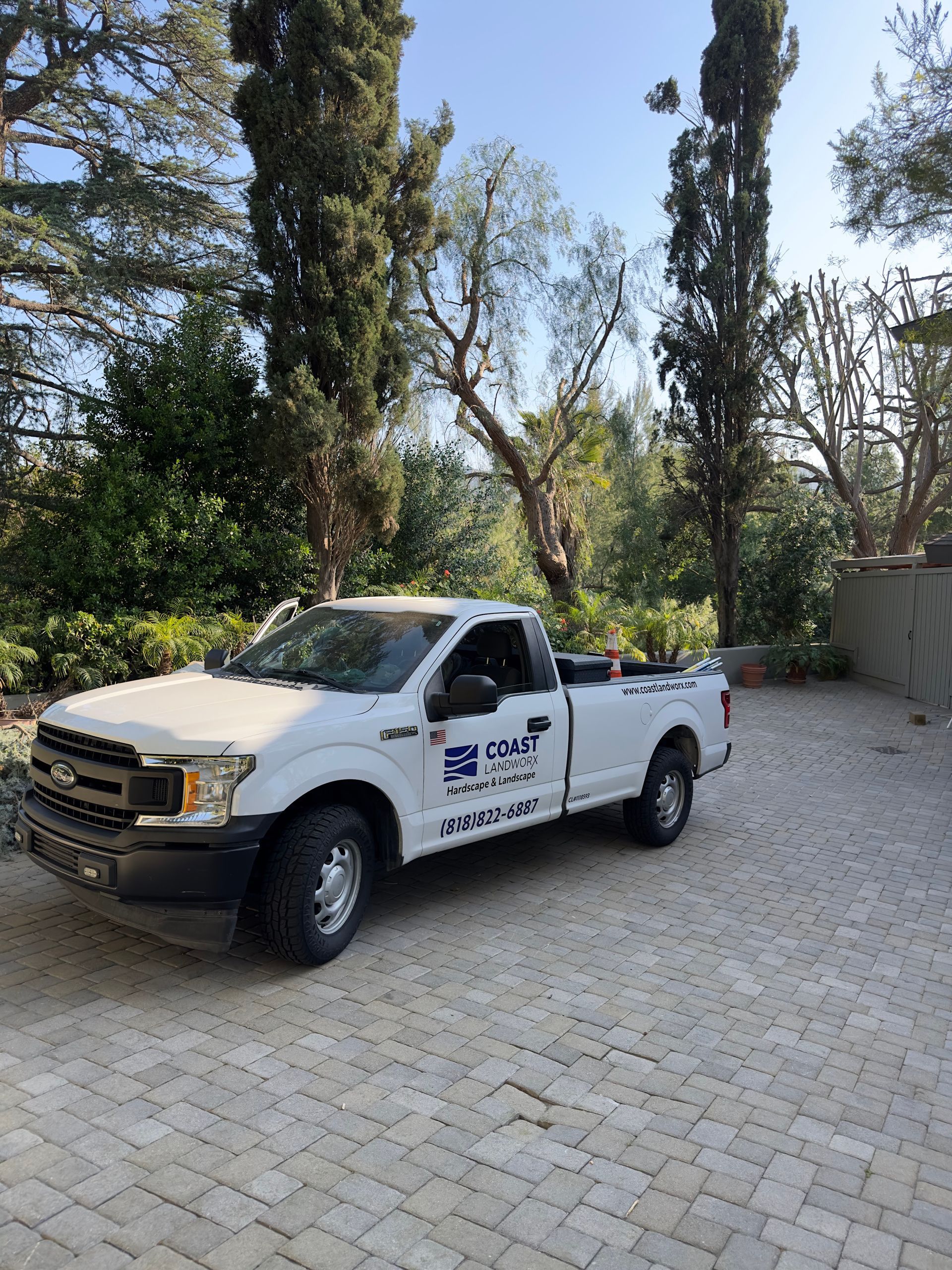 White pickup truck parked on a gravel driveway beside tall trees and a wall