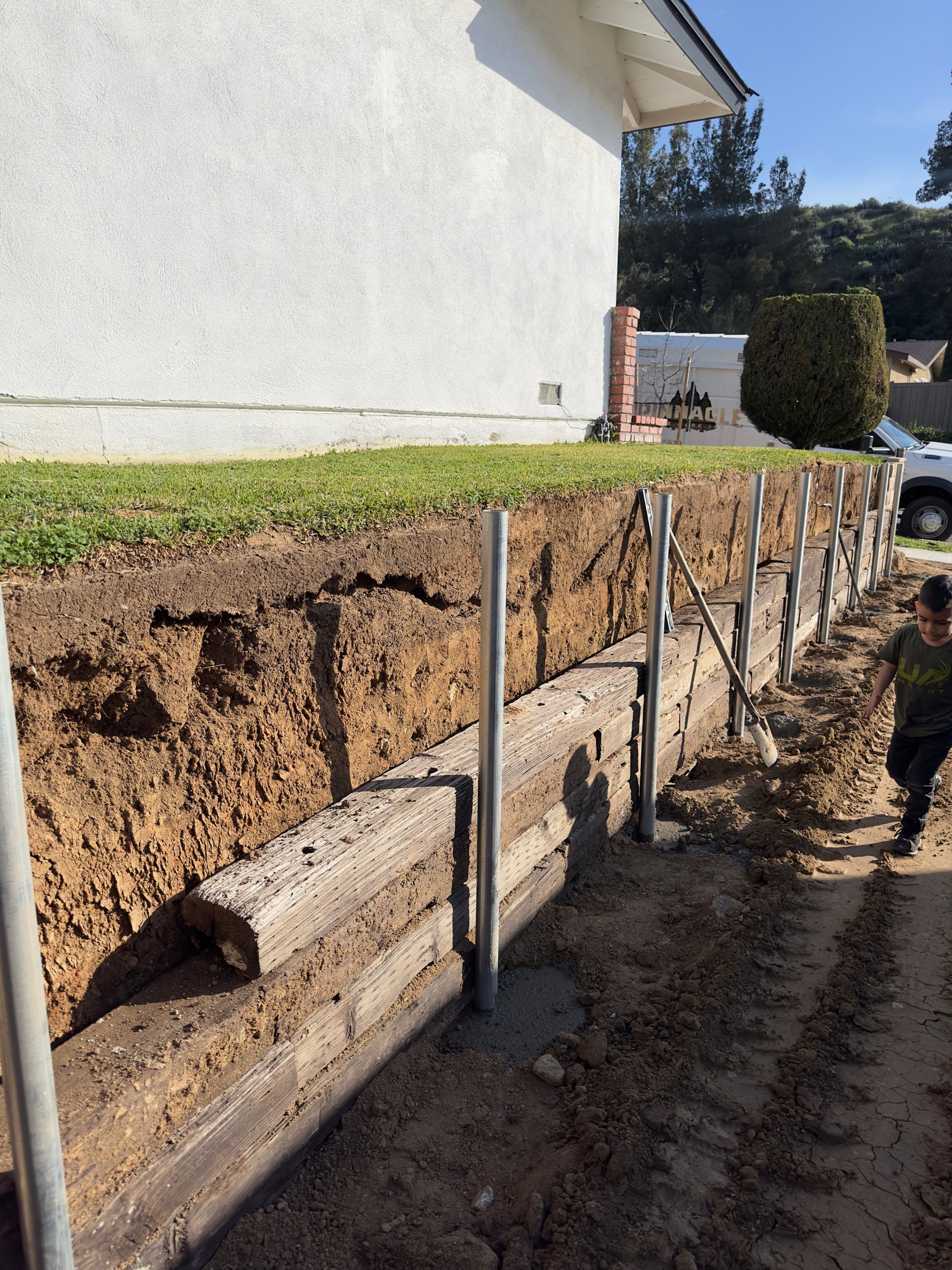 Retaining wall under construction beside a house, with exposed soil and support posts along a sloped yard.
