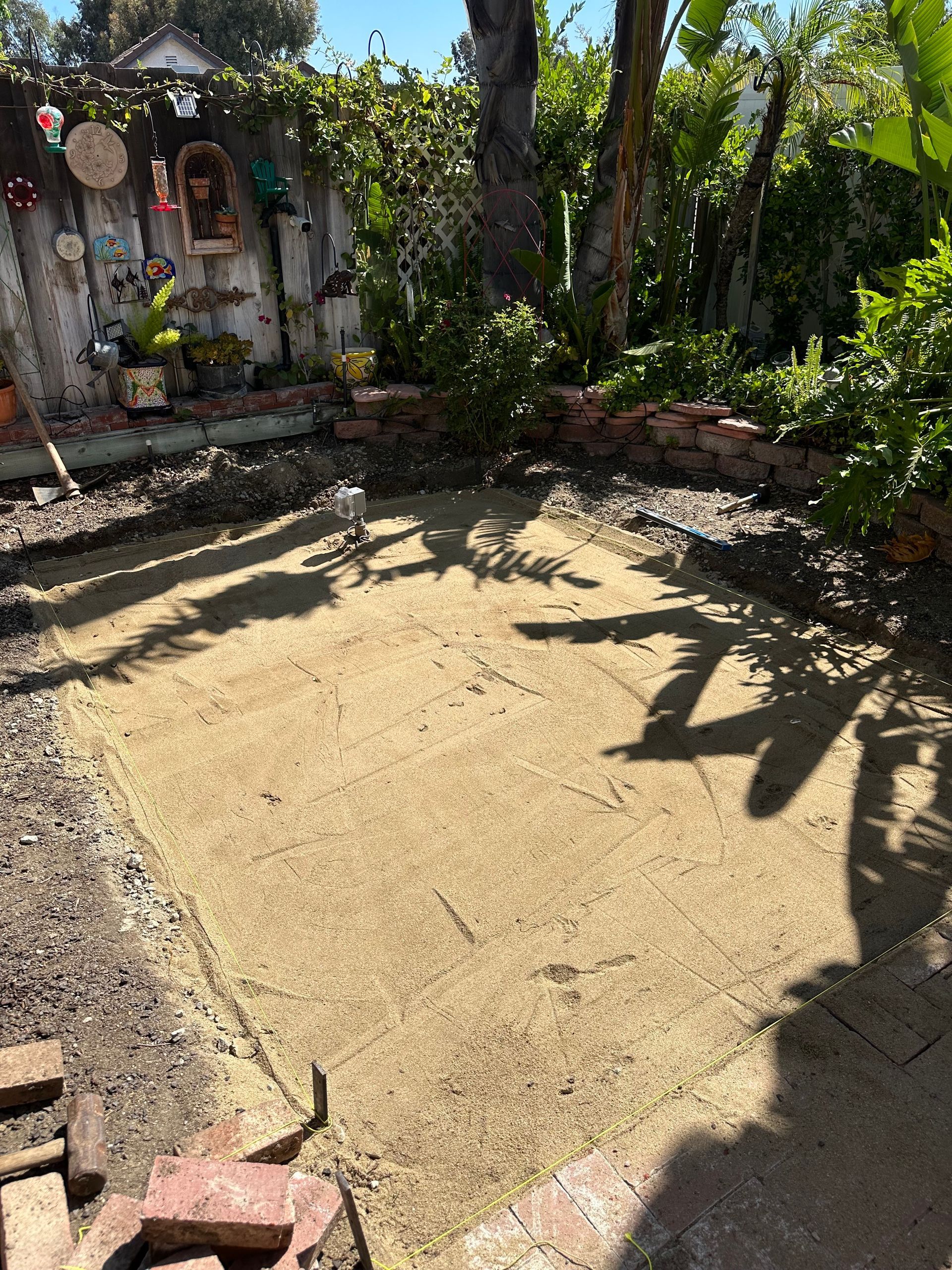Sunny backyard with a small gravel patio, brick border, and lush trees and plants.