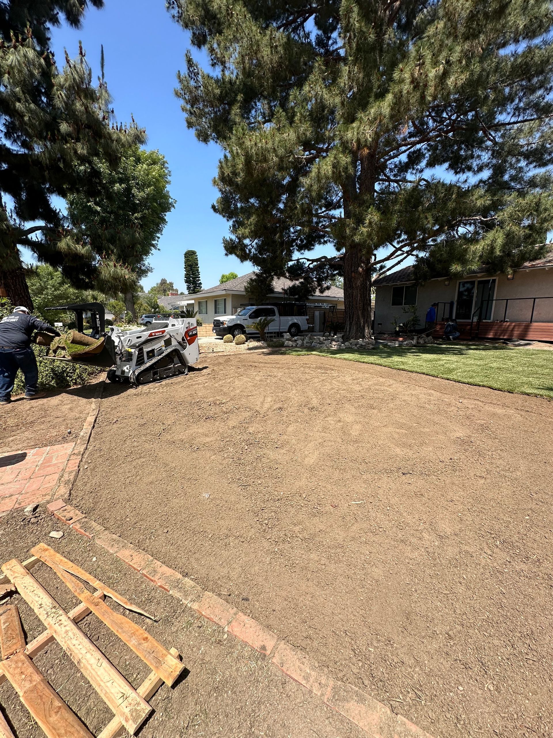 Gravel yard with trees, a house, and a wooden pallet in the foreground under a clear blue sky