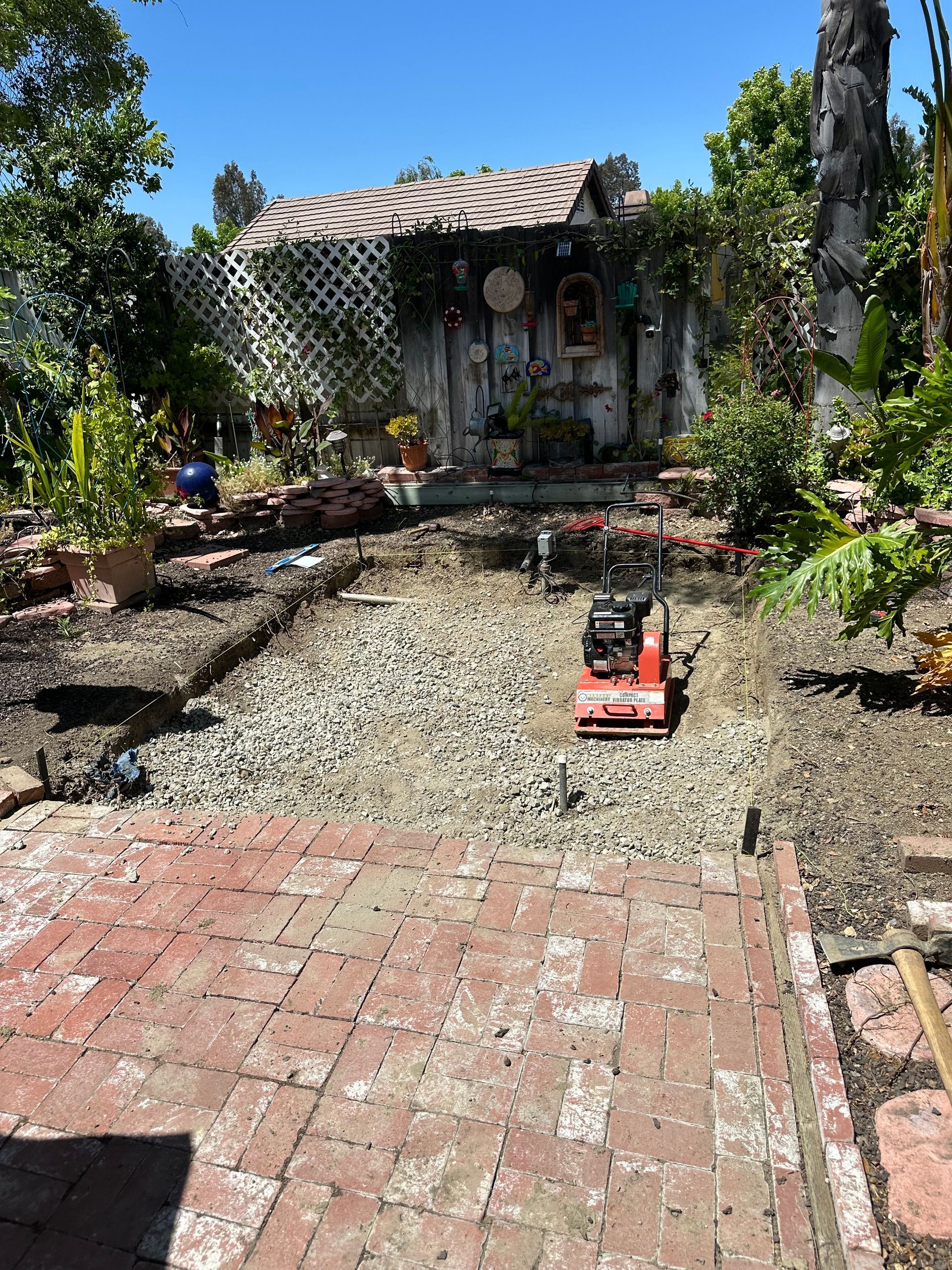 Backyard with red brick patio, gravel path, and a small red mower in front of a house and garden