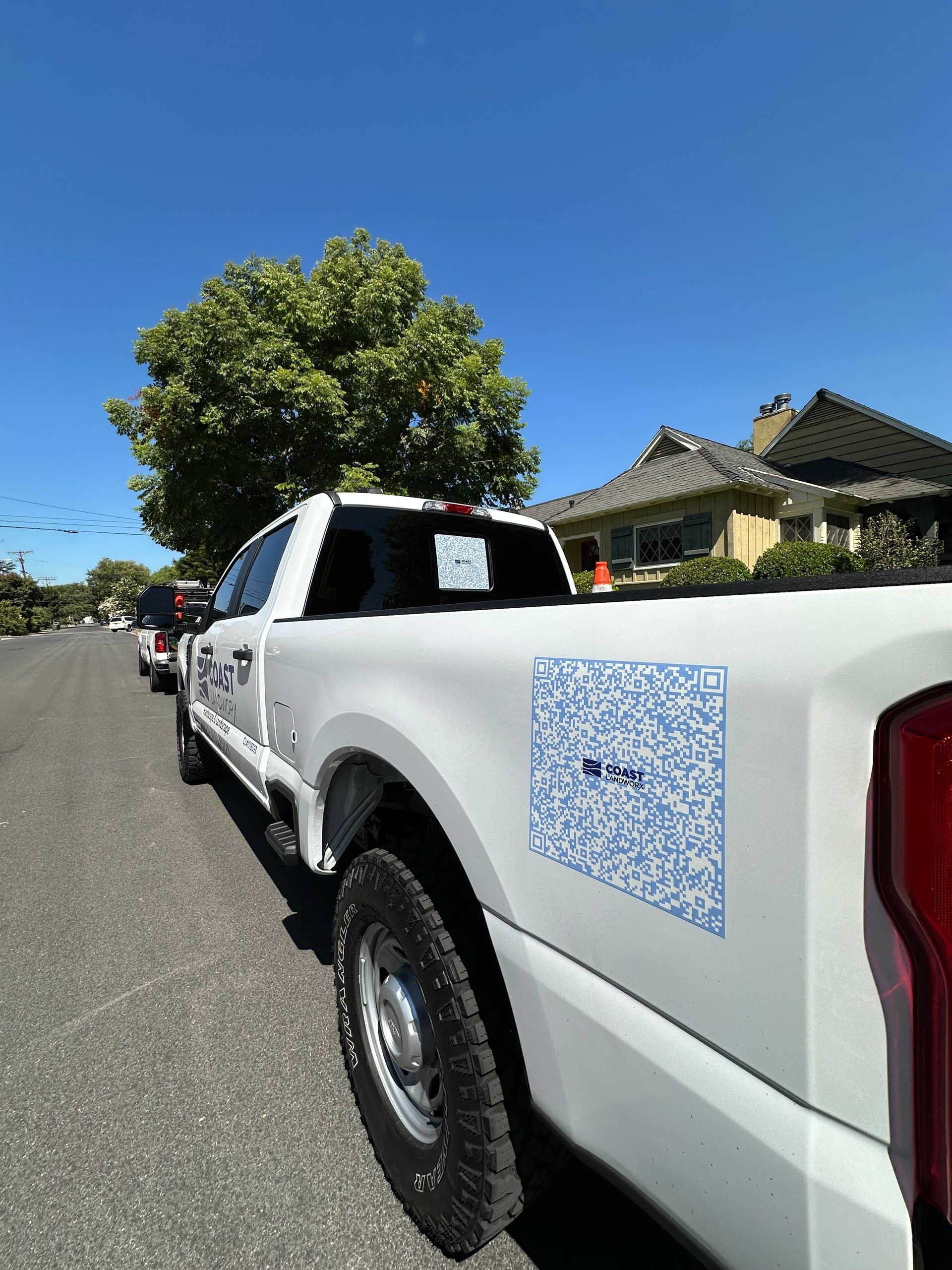 White pickup truck parked on a sunny residential street beside a house and a leafy tree
