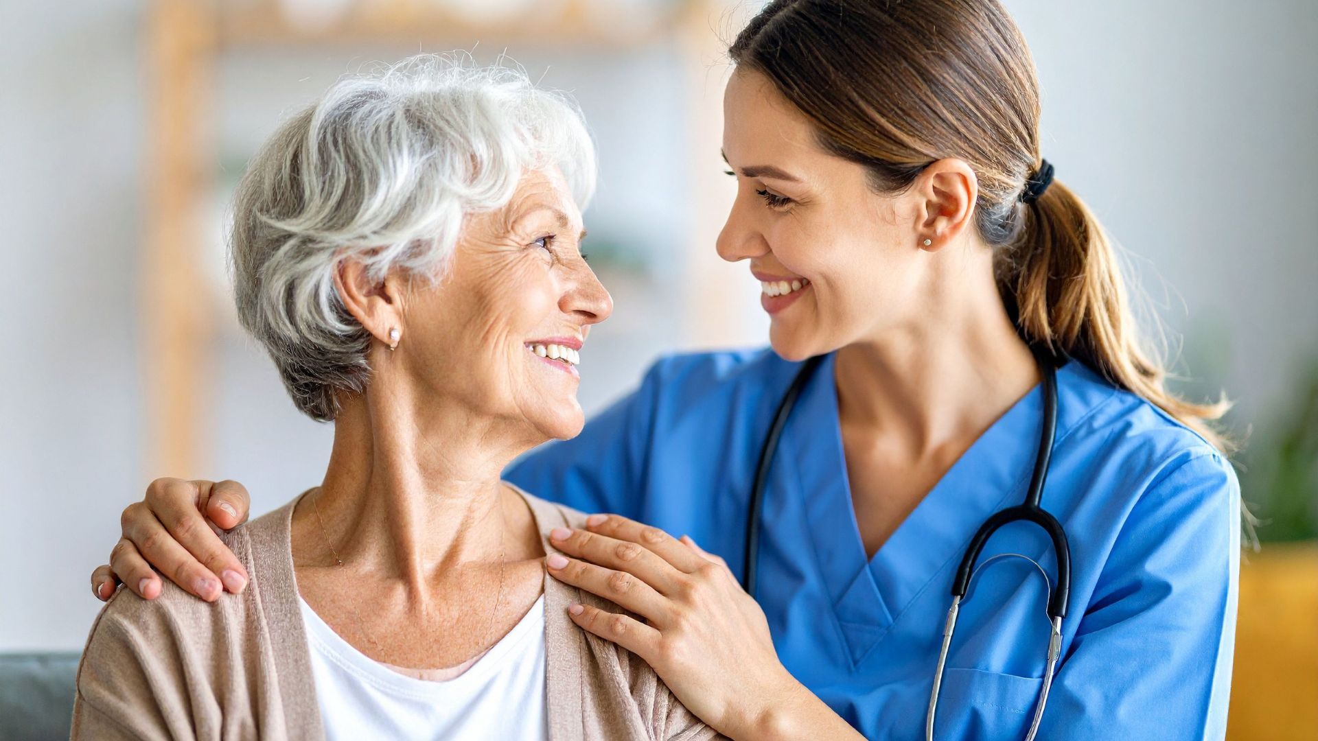 Nurse in blue scrubs comforting an elderly woman, smiling face to face in a bright room