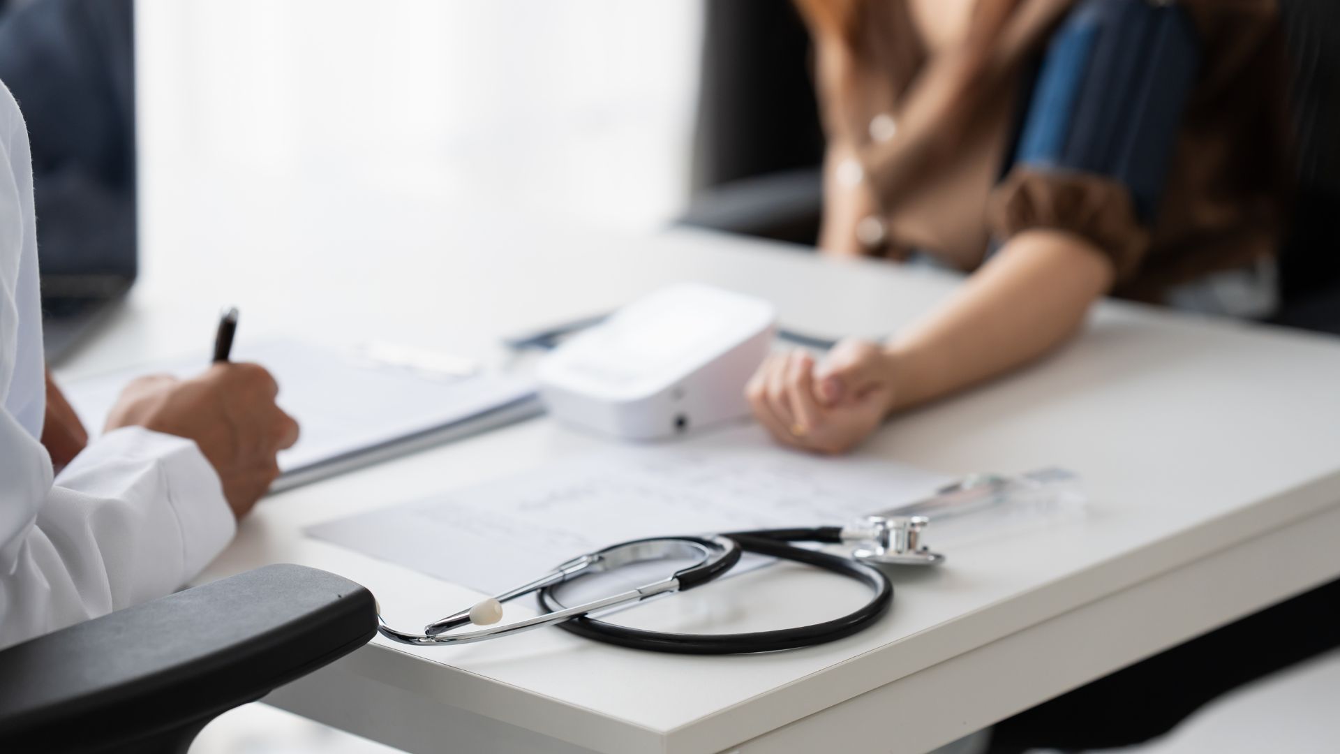 Doctor’s office desk with stethoscope, clipboard, and a patient in discussion across the table