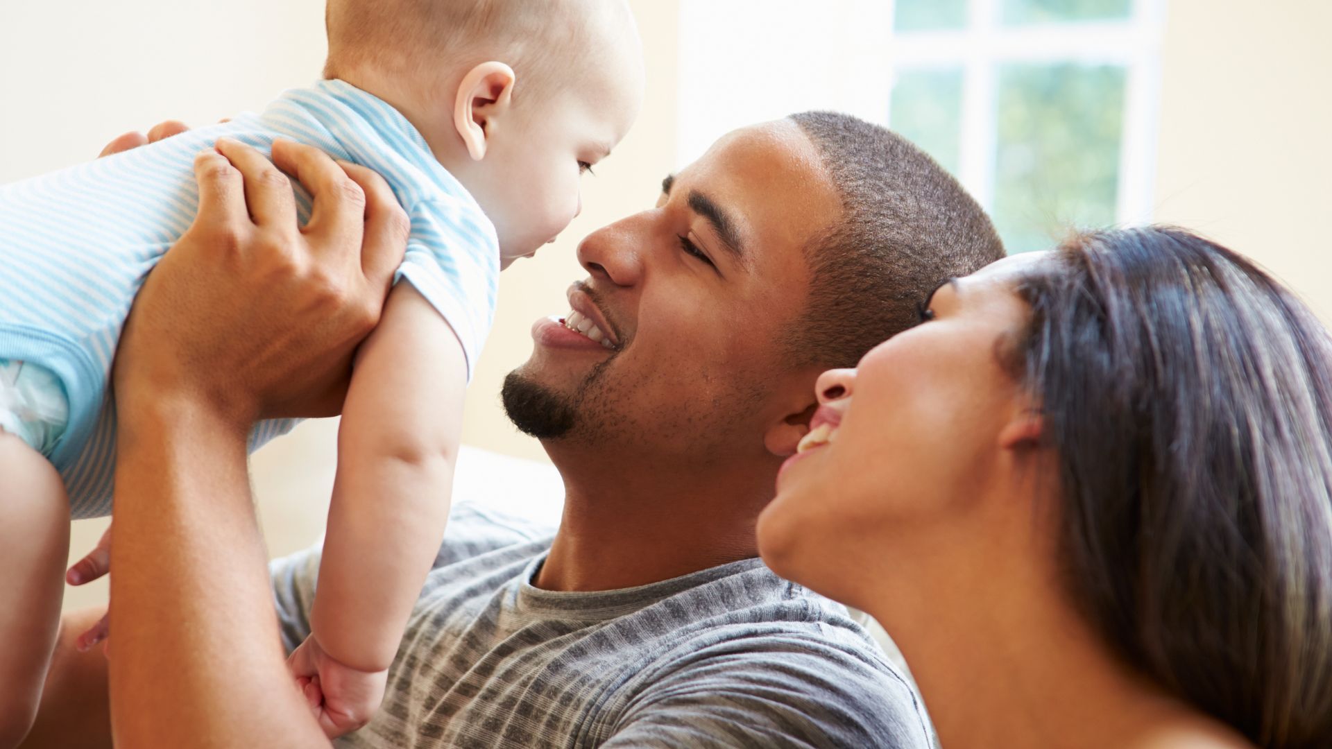 A smiling parent lifting a baby while another parent looks on in a bright room.