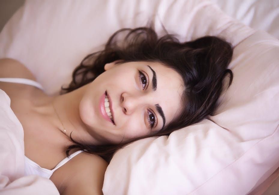 Woman reclining on a bed, smiling softly on white pillows with dark hair spread around her.