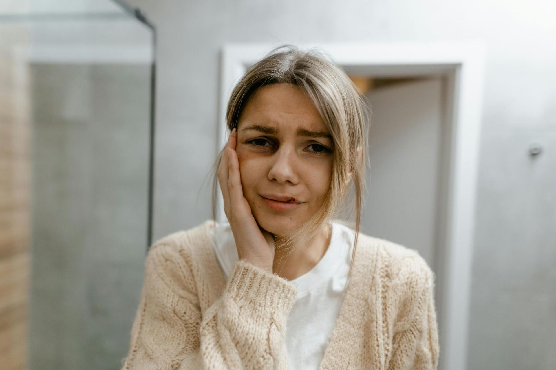 Woman in a beige sweater touching her face, looking worried in a bathroom mirror.