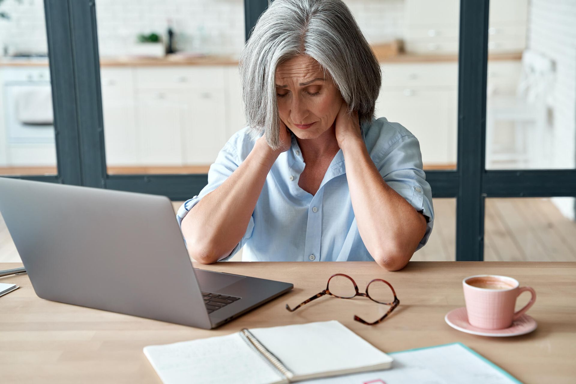 Person sitting at a desk with a laptop, holding their head in a bright home office